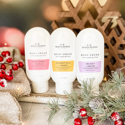 Three bottles of 'Simply Stated' body crème with holiday decorations in the background