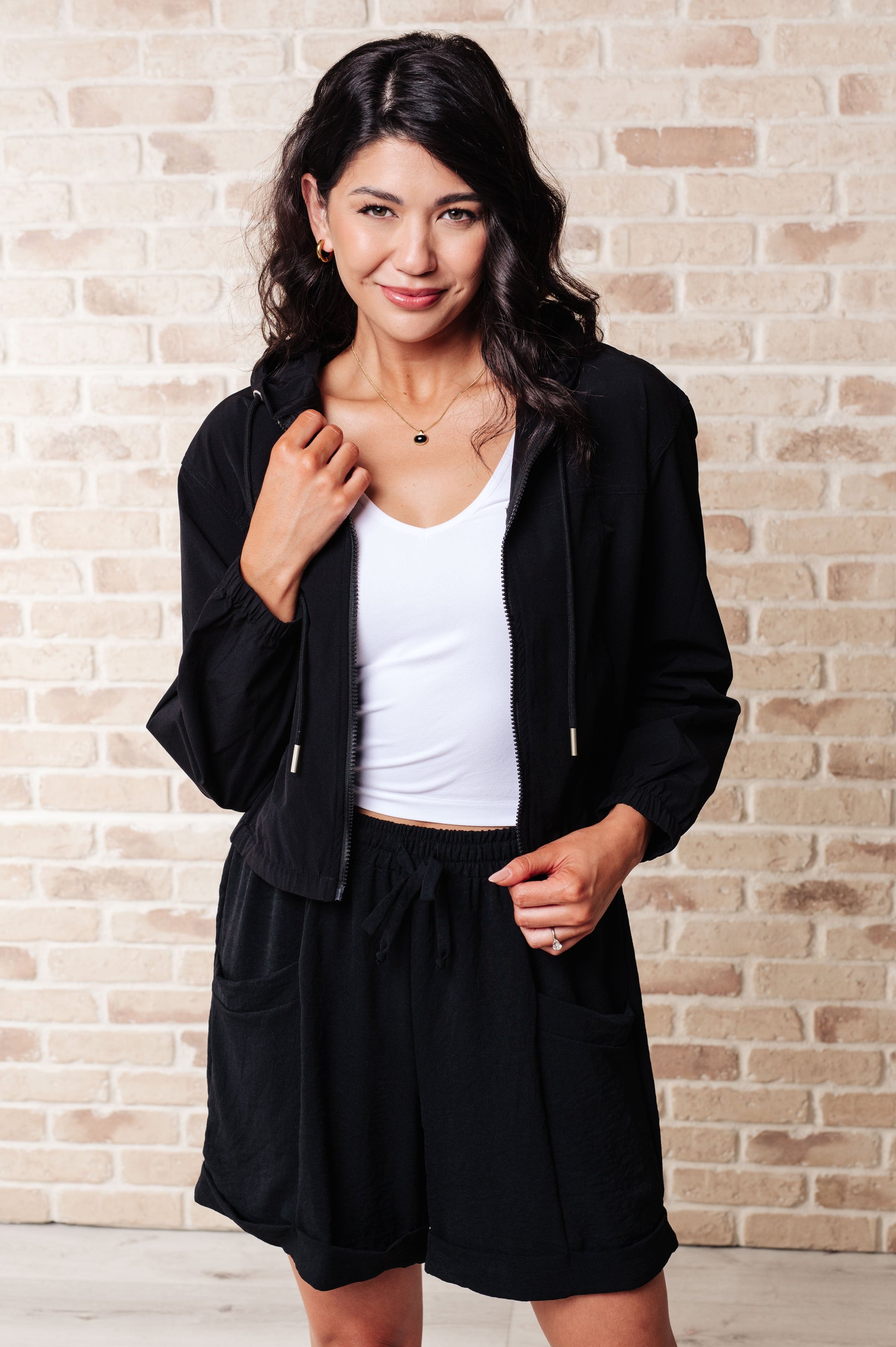 Woman wearing a black zip up jacket and shorts with a white top against a brick wall.