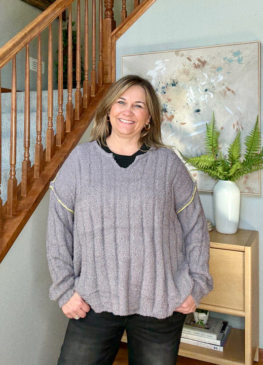 Woman wearing a gray sweater standing in a home setting with a staircase and artwork in the background.