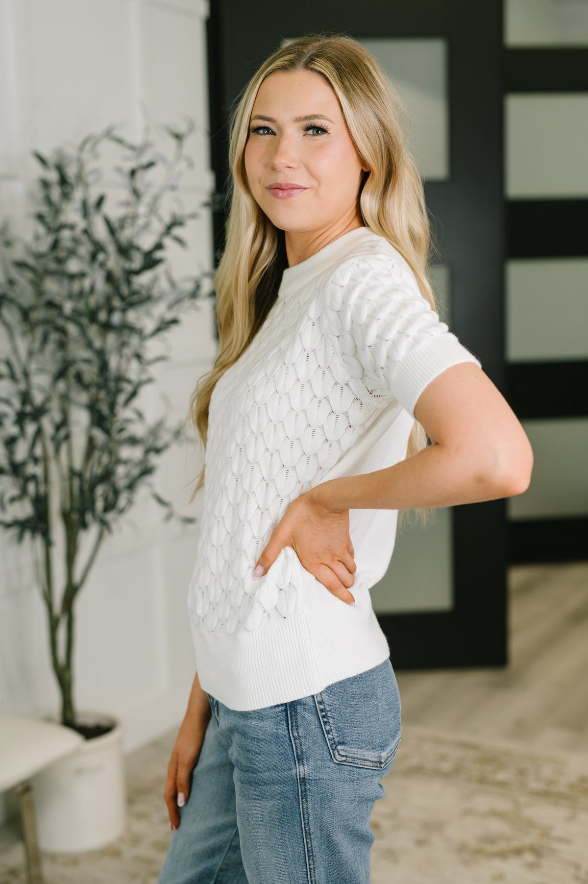 A woman modeling a white short-sleeve knit sweater with a textured cable pattern, paired with blue jeans in a modern indoor setting.