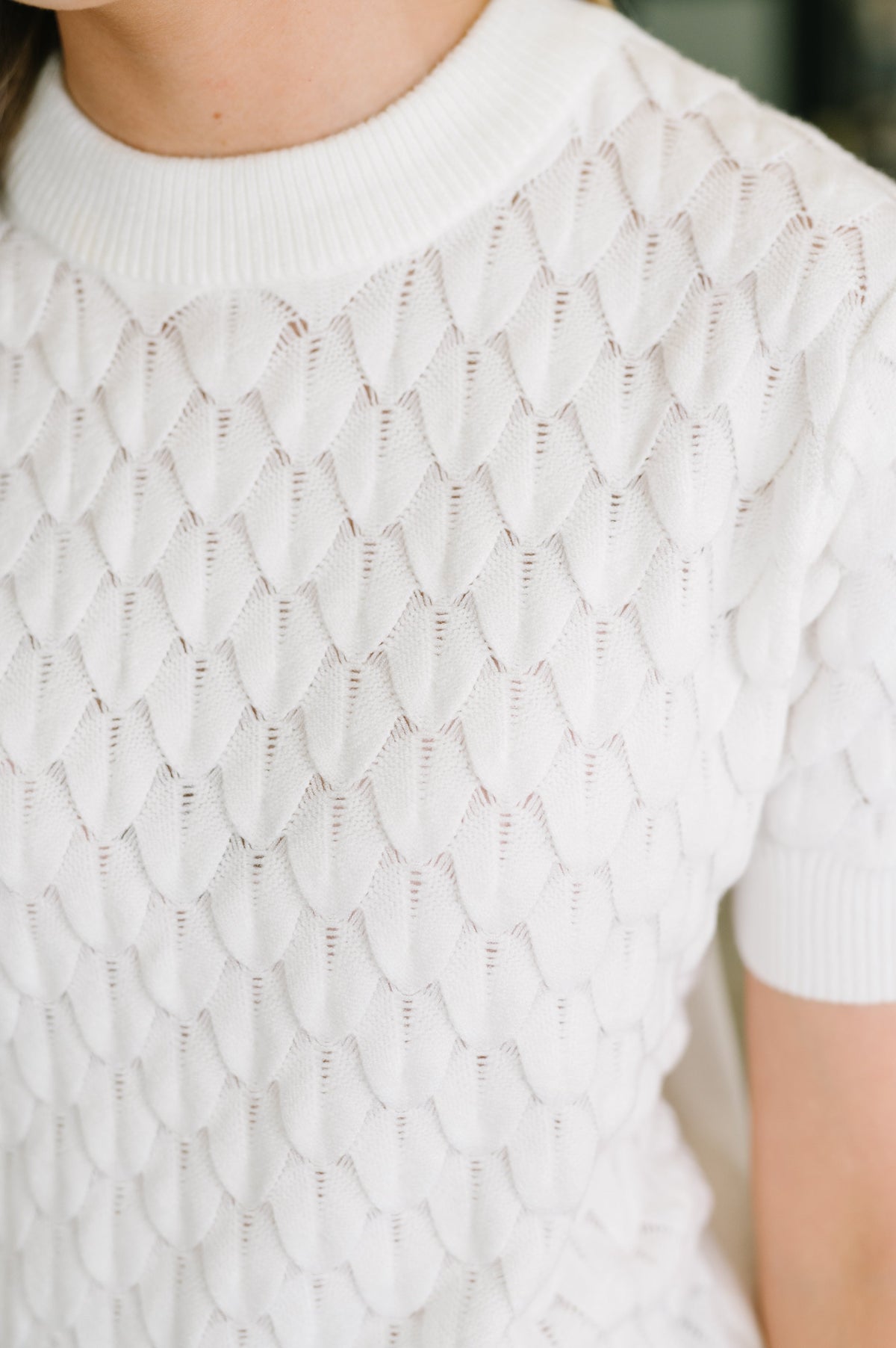 Close-up of a person wearing a white short-sleeve knit top with an openwork scalloped or fish-scale pattern across the chest and shoulders.