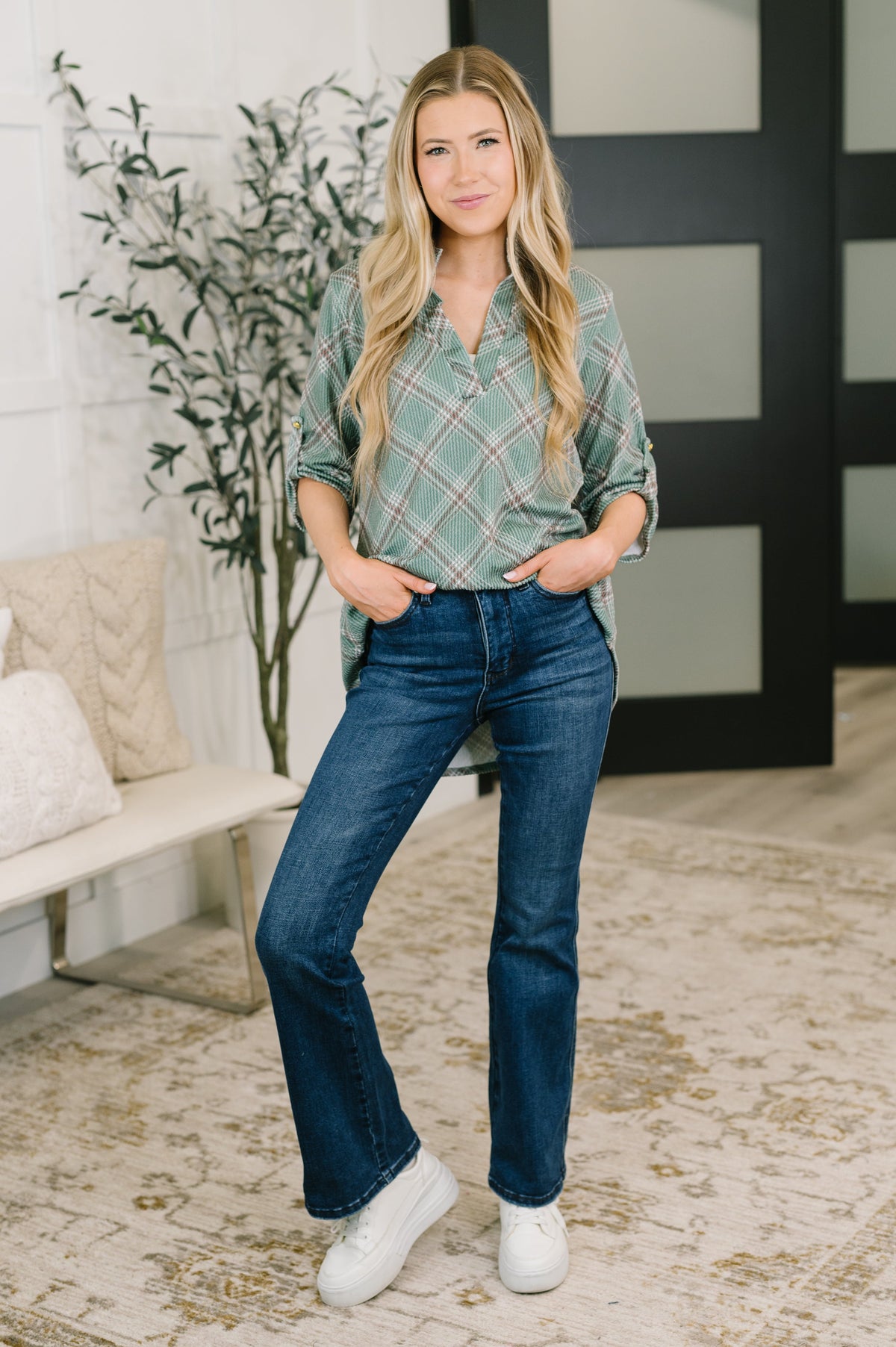 A woman modeling a green and white plaid button-down shirt with dark blue flare jeans and white sneakers, standing in a modern indoor setting with a bench, plant, and patterned rug.