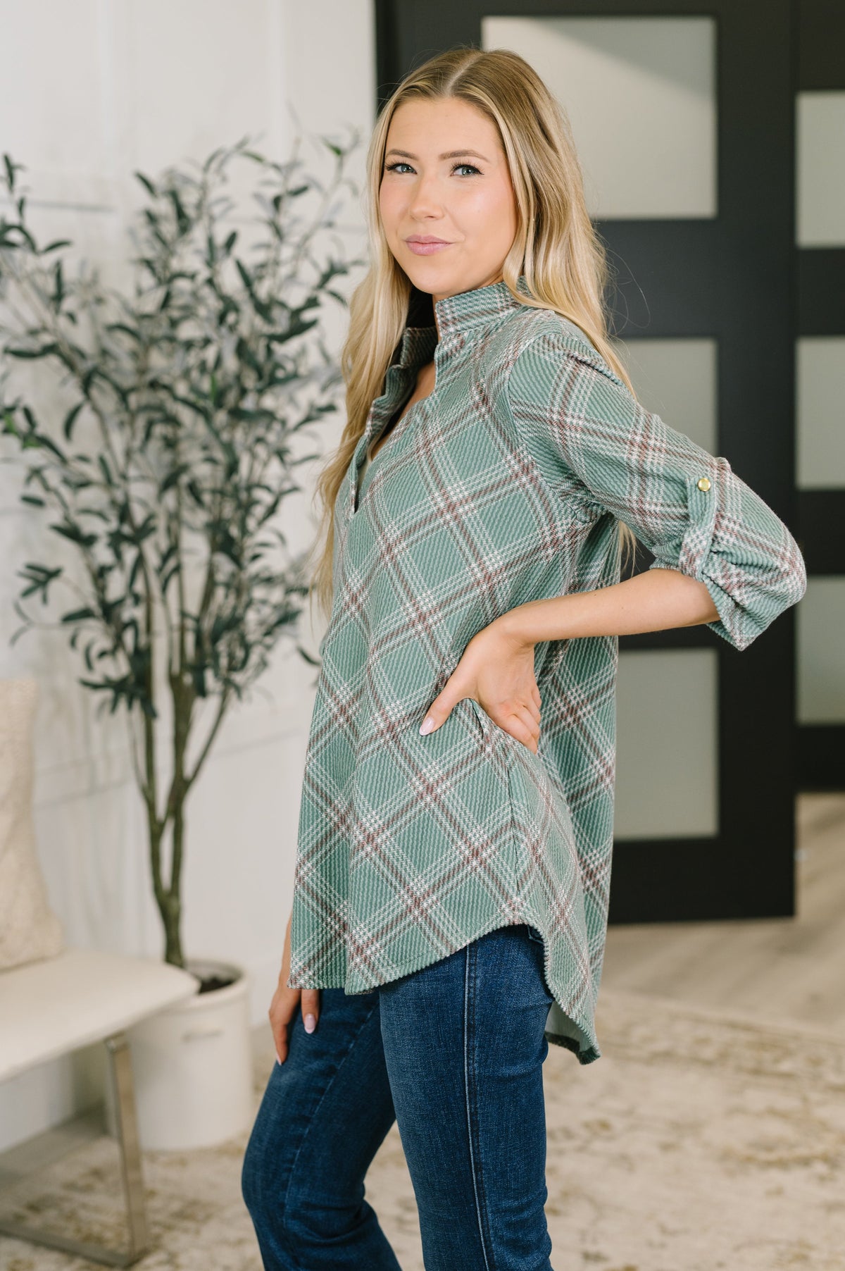 A woman modeling a sage green plaid button-up shirt with rolled-up sleeves and dark blue jeans, standing in a modern indoor setting with a potted plant in the background.