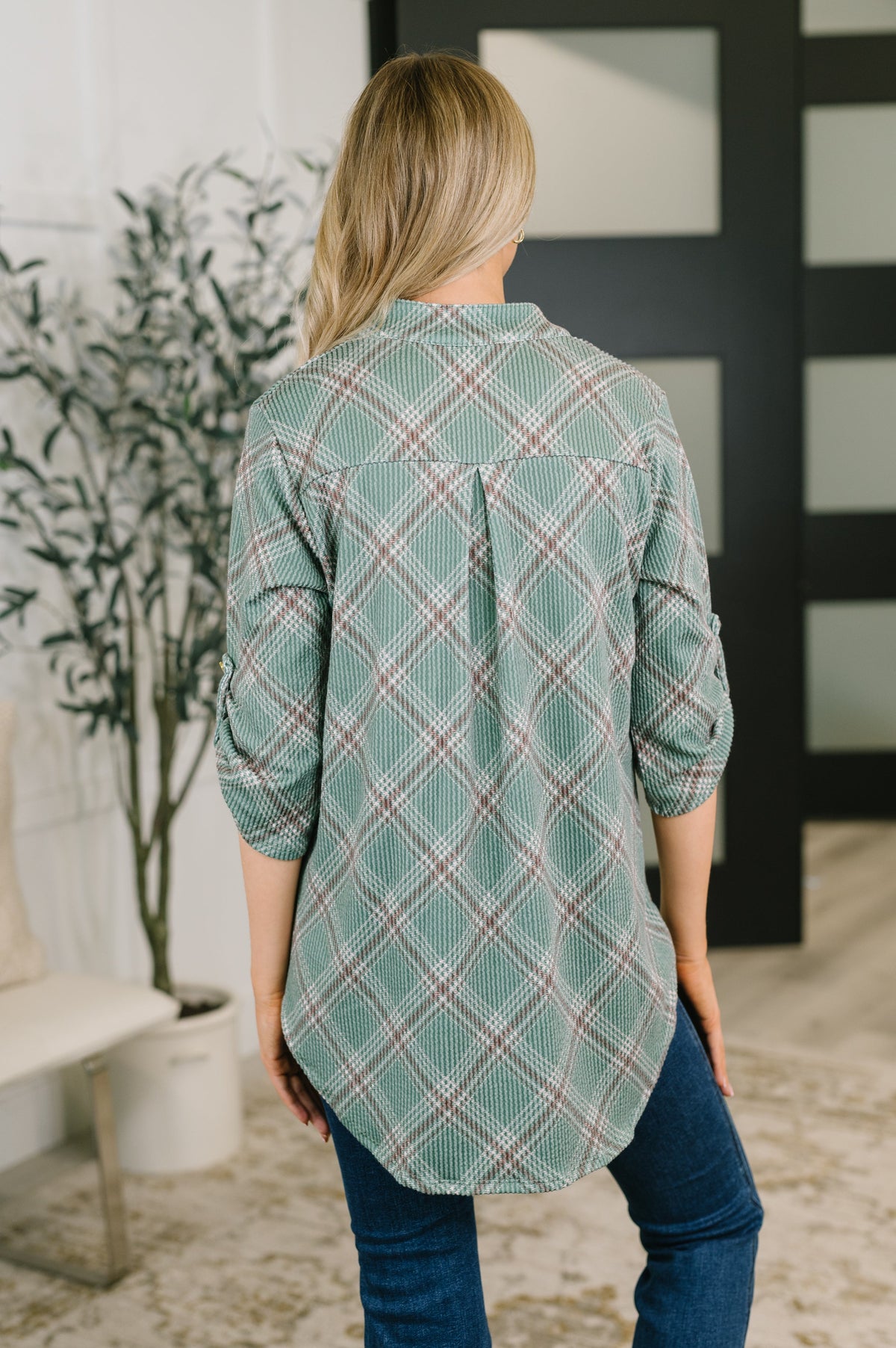 A woman viewed from behind wearing a sage green plaid button-up shirt with rolled-up sleeves and blue jeans in a modern indoor setting.