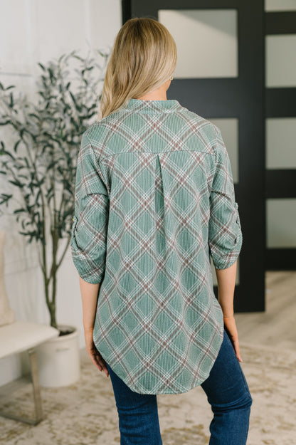 A woman viewed from behind wearing a sage green plaid button-up shirt with rolled-up sleeves and blue jeans in a modern indoor setting.