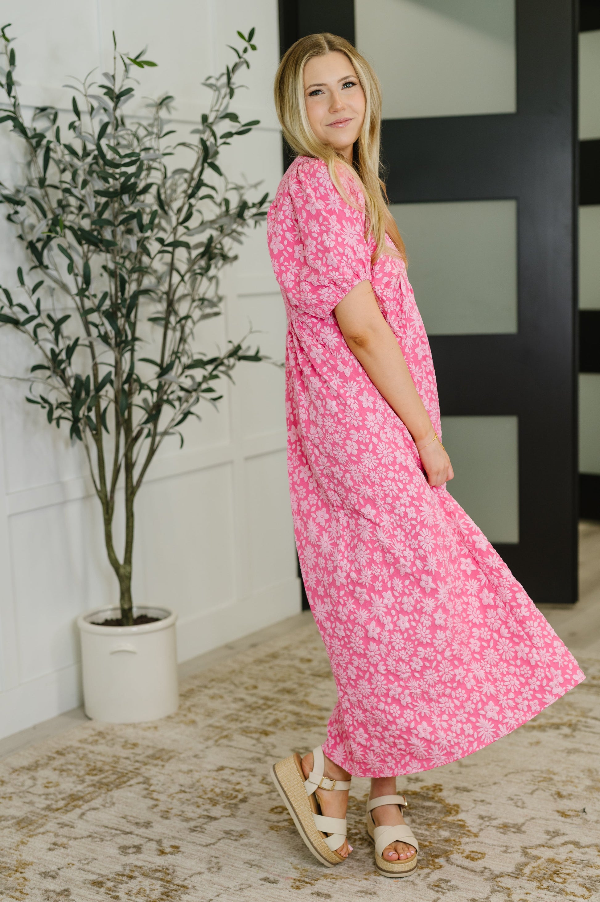 Woman in a pink floral dress standing indoors next to a plant.