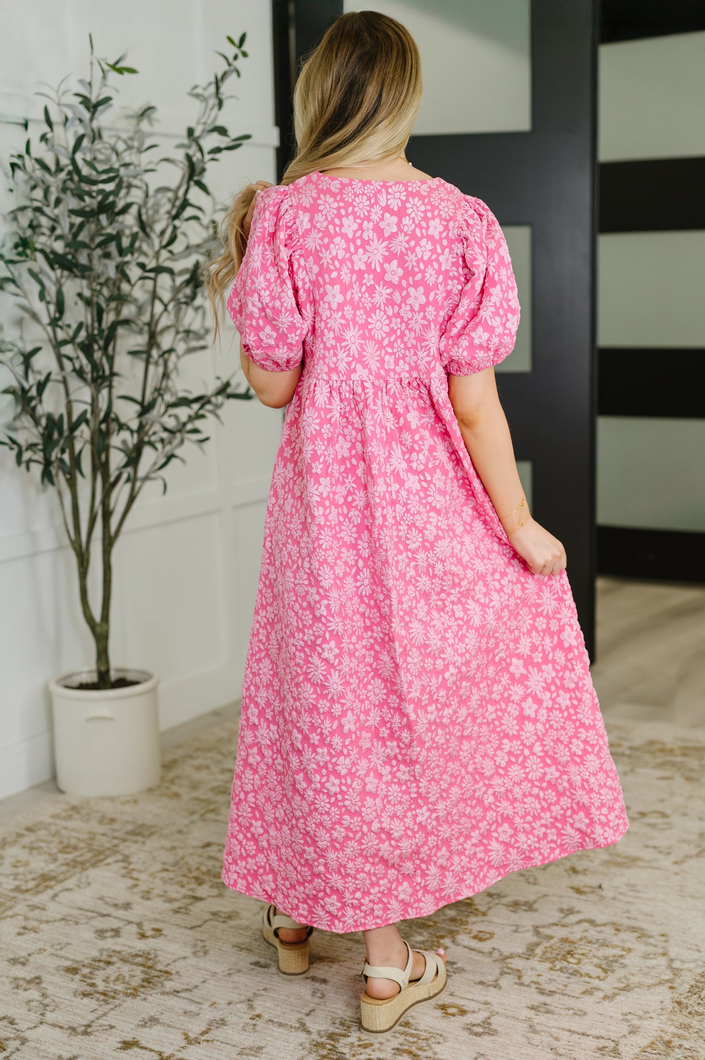 Woman wearing a pink floral dress standing in a room with a plant and striped wall.