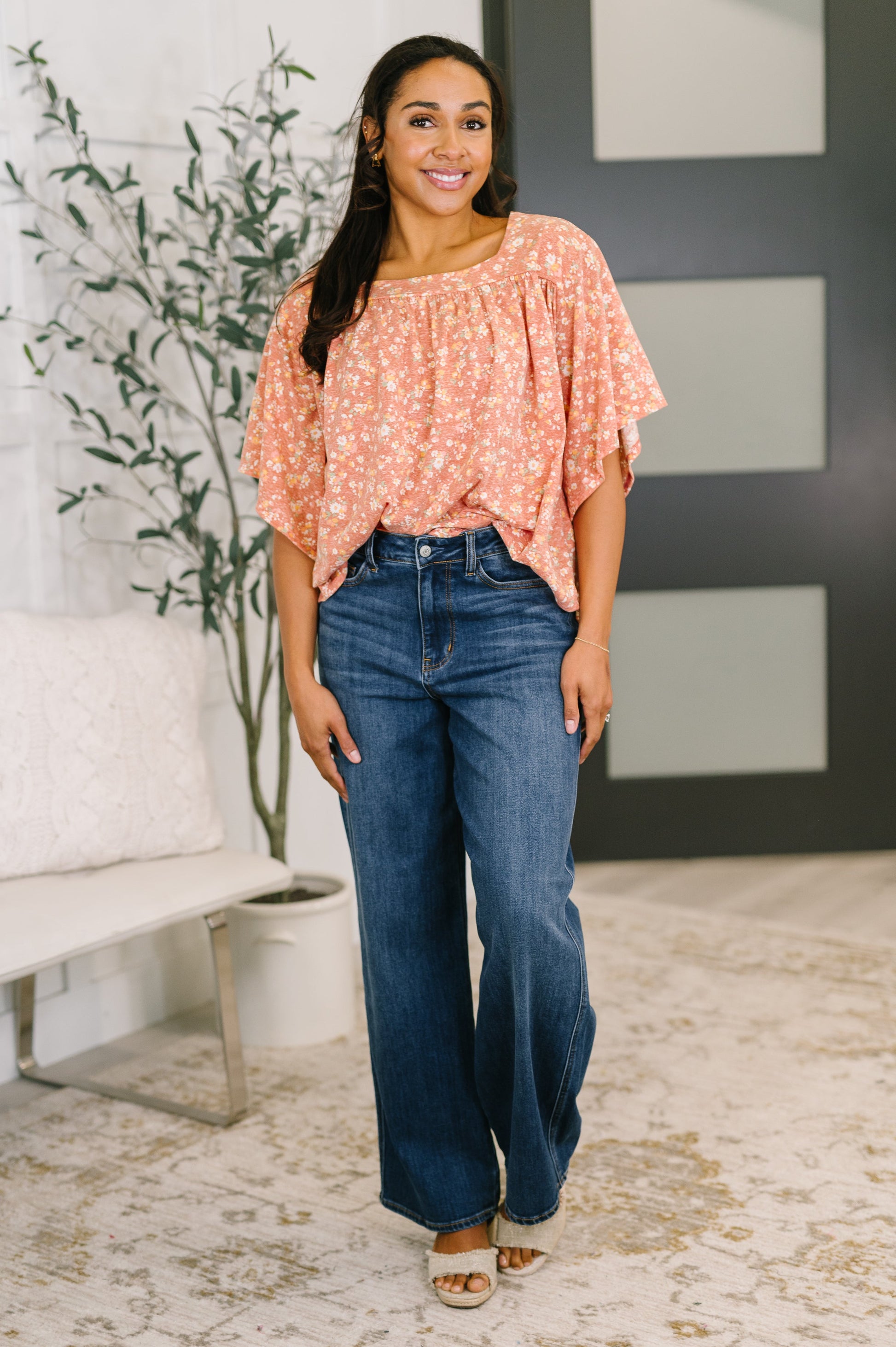 Woman wearing a peach floral top and blue jeans standing in a room with a plant and white couch.