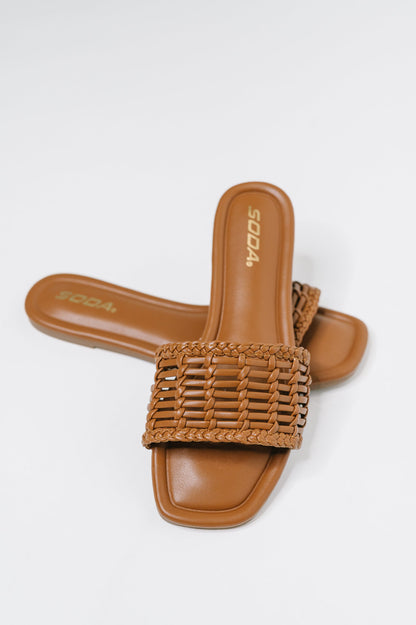 A pair of tan slide sandals with a woven strap, shown on a light gray background. The brand name 'SODA' is visible on the insole.