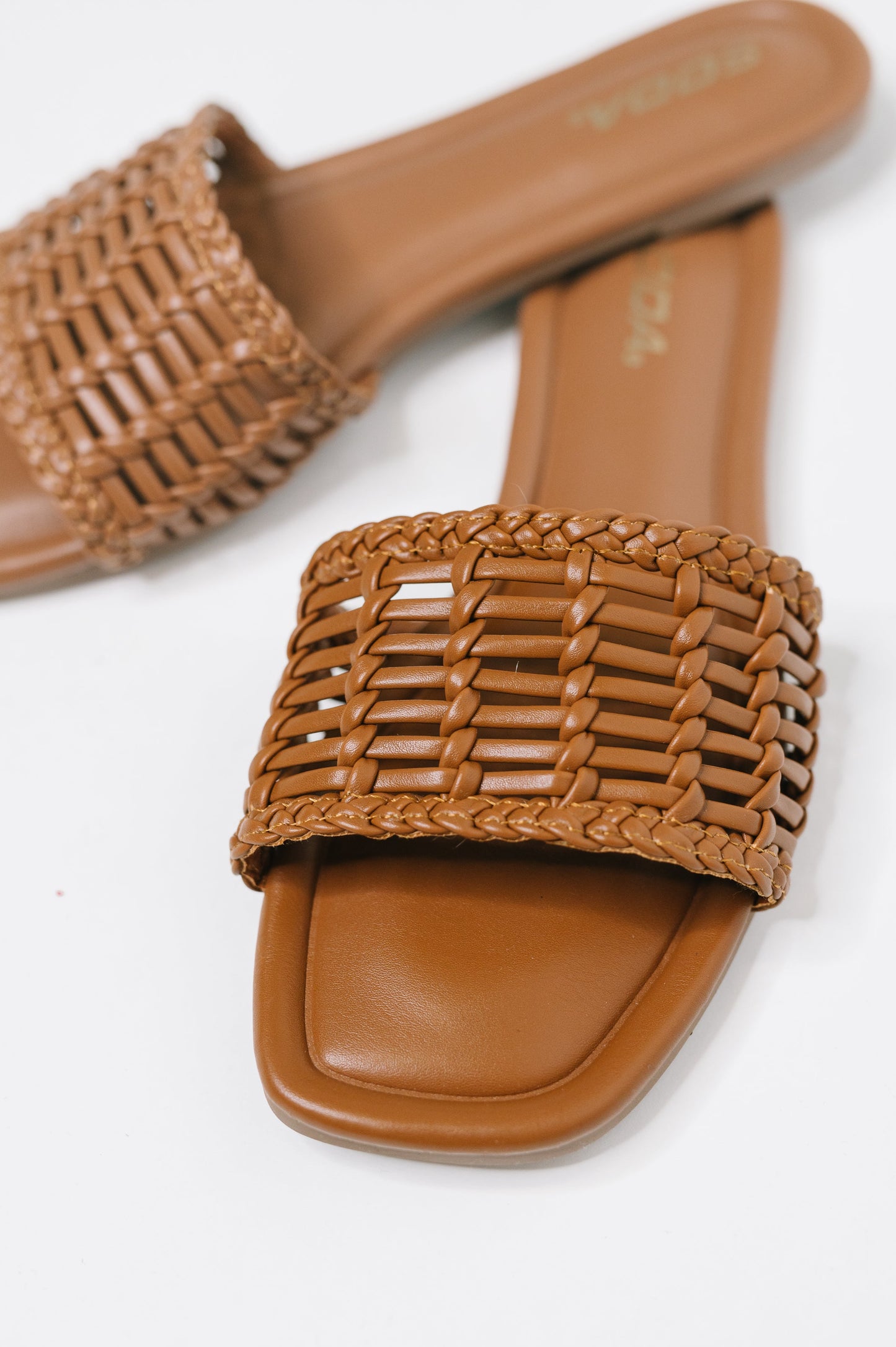 A pair of tan woven slide sandals with square toes, featuring a braided upper strap and a smooth tan footbed, shown on a white background.
