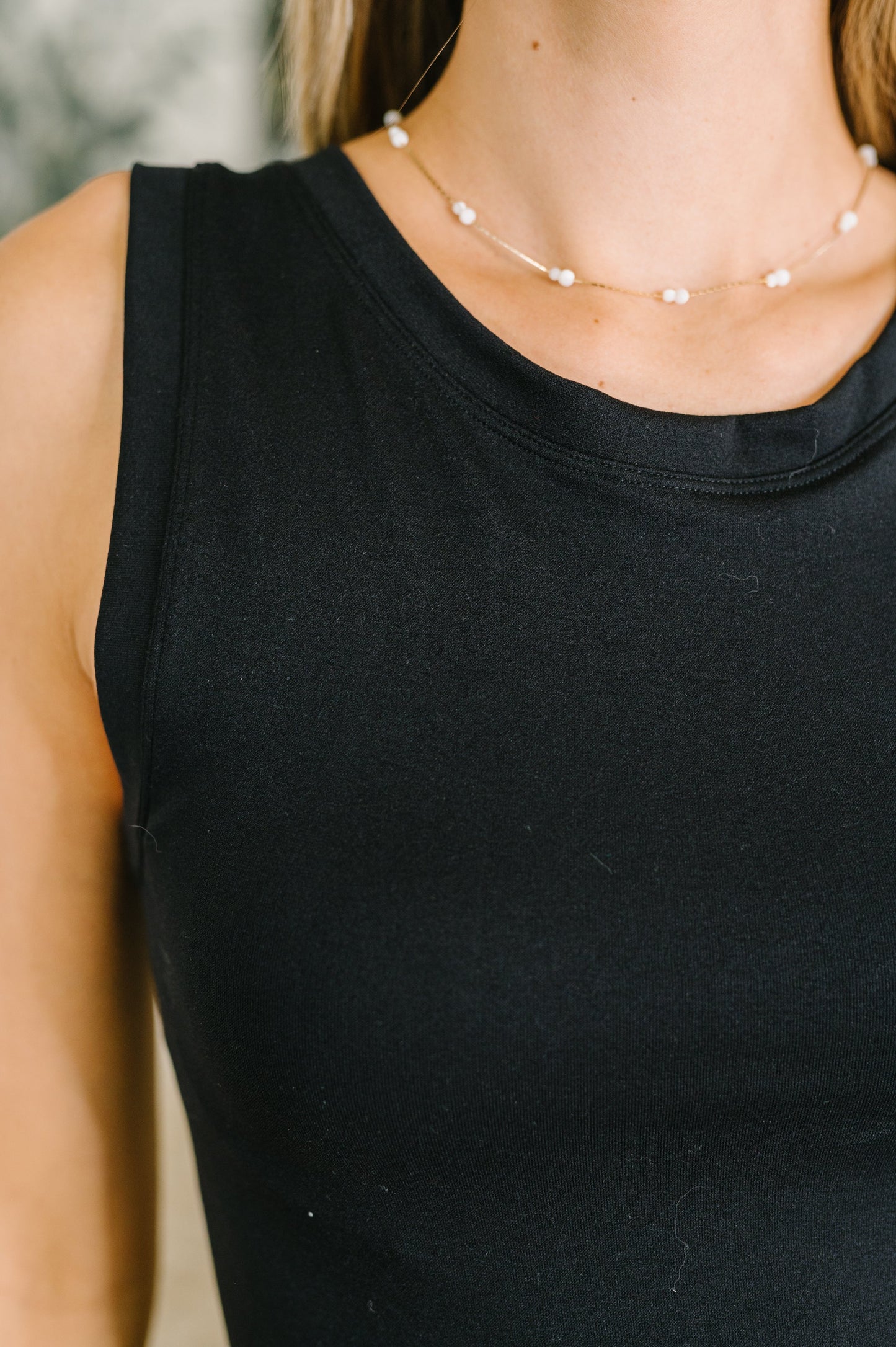 Person wearing a black sleeveless top and pearl necklace against a blurred background