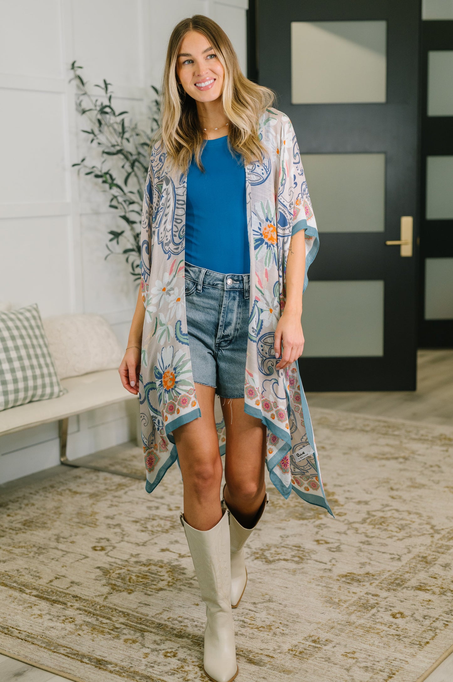 Woman wearing a floral kimono over a blue top and denim shorts, standing in a room with a couch and plant.