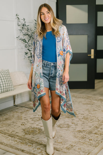 Woman wearing a floral kimono over a blue top and denim shorts, standing in a room with a couch and plant.