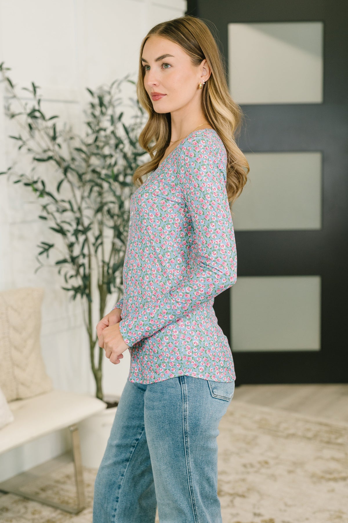 A woman modeling a long-sleeve V-neck top with a dense small floral print in pink, blue, and green, paired with light-wash blue jeans in a modern indoor setting.