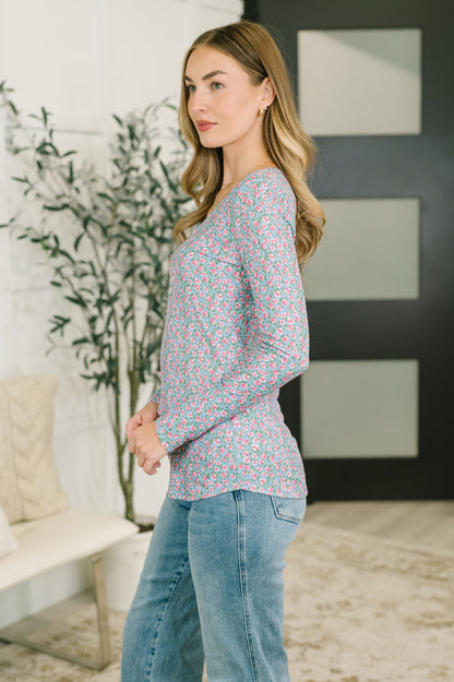 A woman modeling a long-sleeve V-neck top with a dense small floral print in pink, blue, and green, paired with light-wash blue jeans in a modern indoor setting.