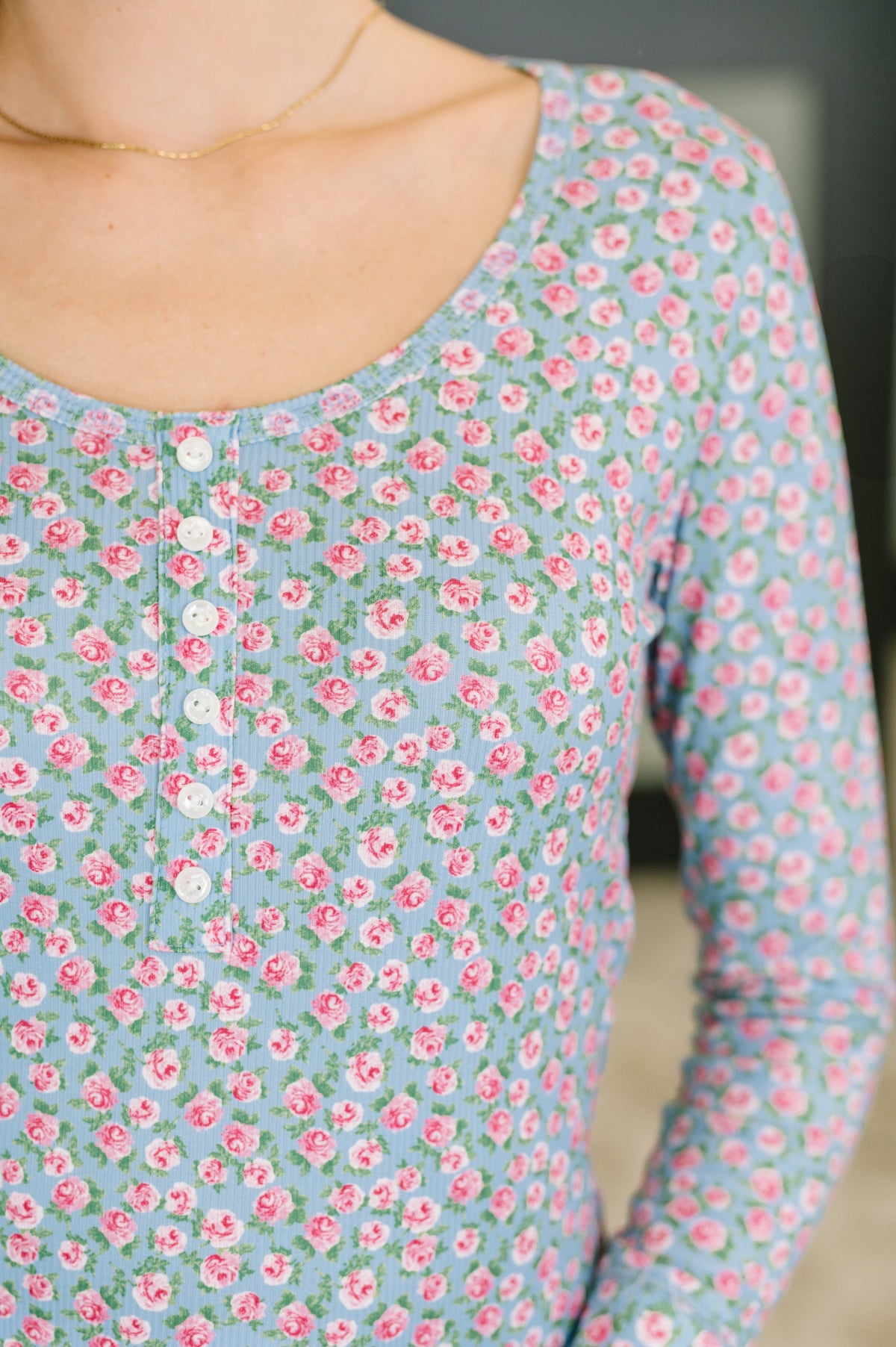 A close-up of a person wearing a light blue long-sleeve top with a pink and green floral print, featuring a buttoned henley-style neckline.
