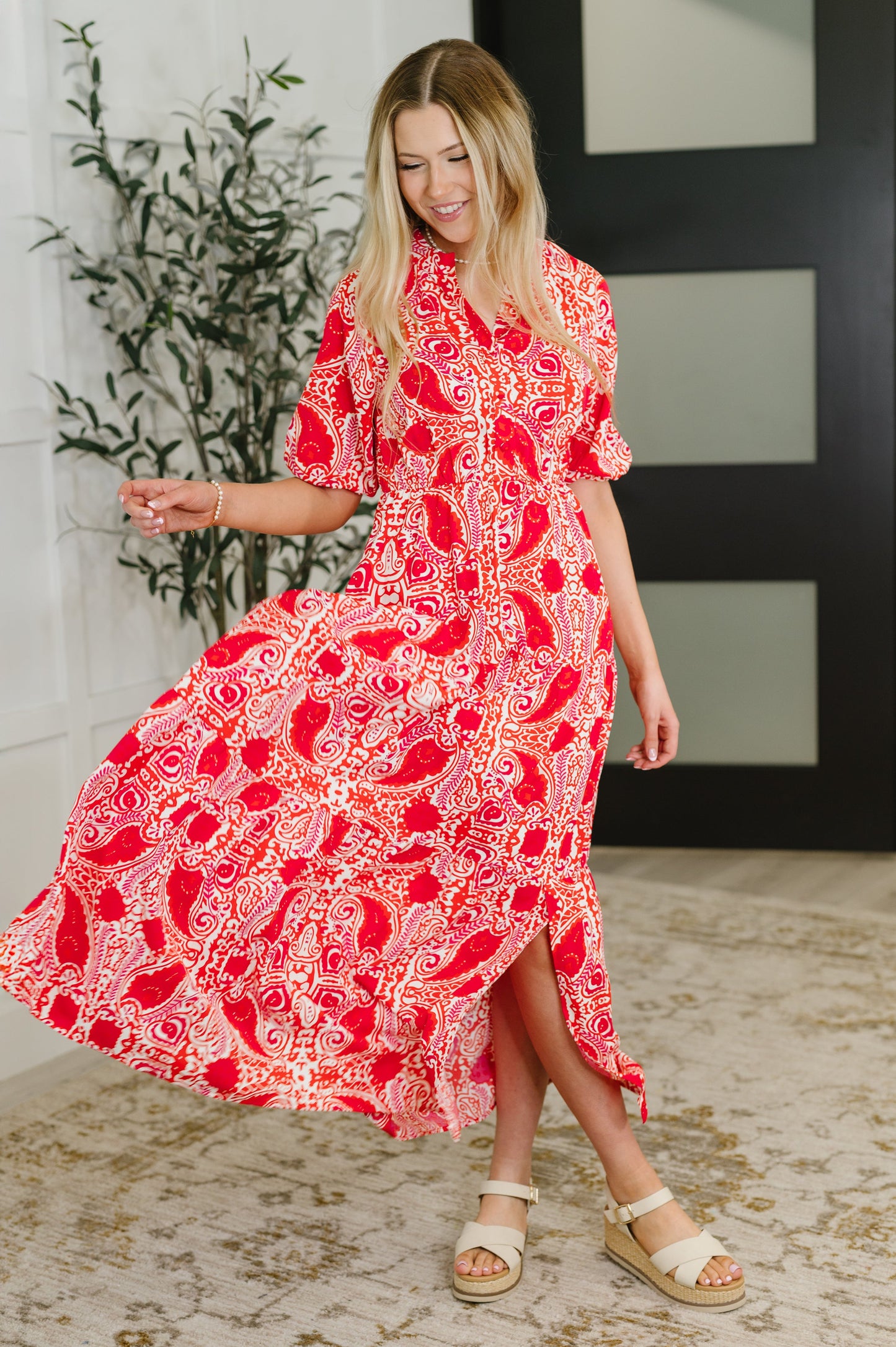 Woman wearing a red patterned dress standing indoors with a plant and wall decor in the background.