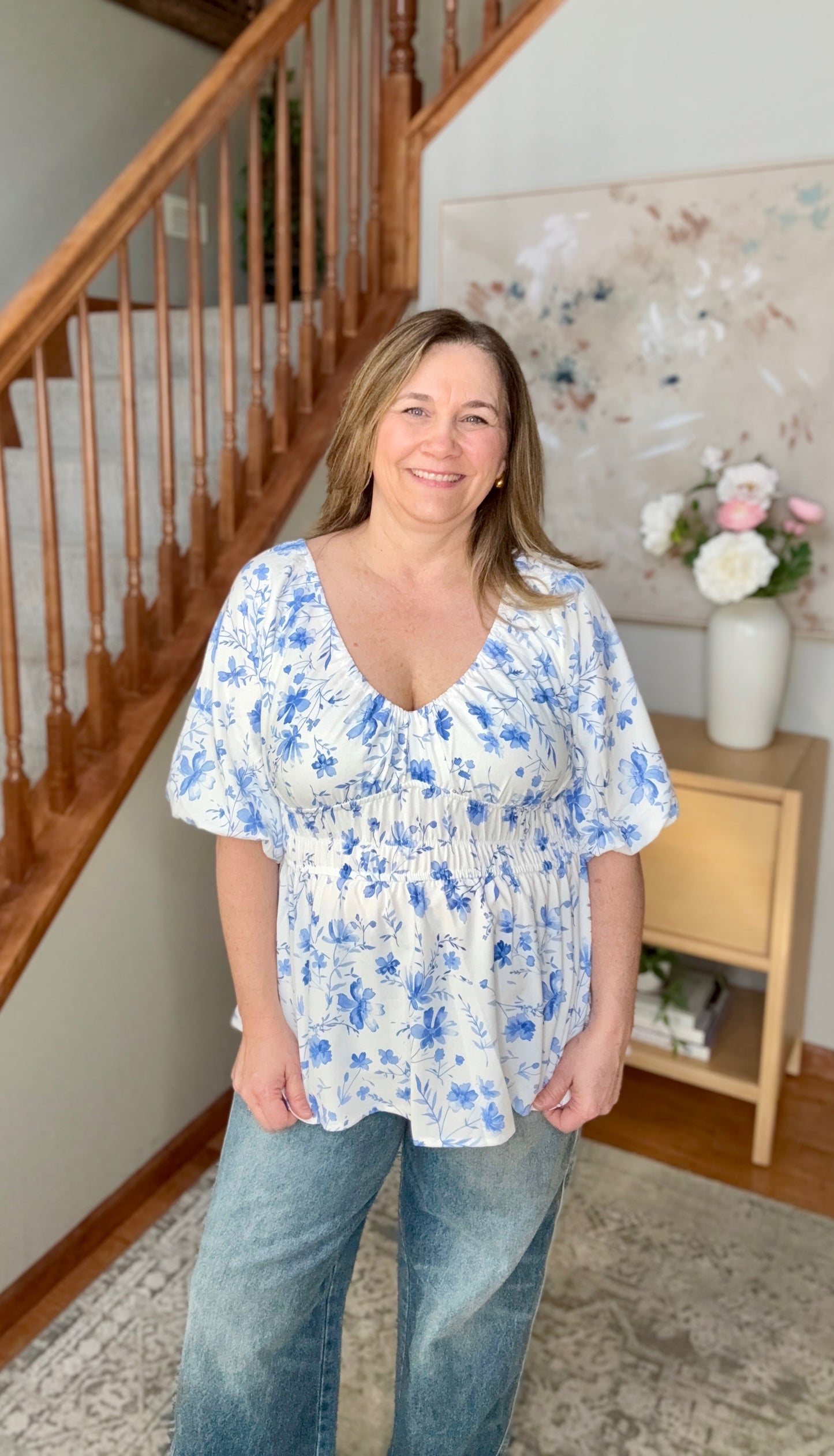 Woman wearing a blue floral blouse and jeans standing in a home setting with a staircase and decorative items.