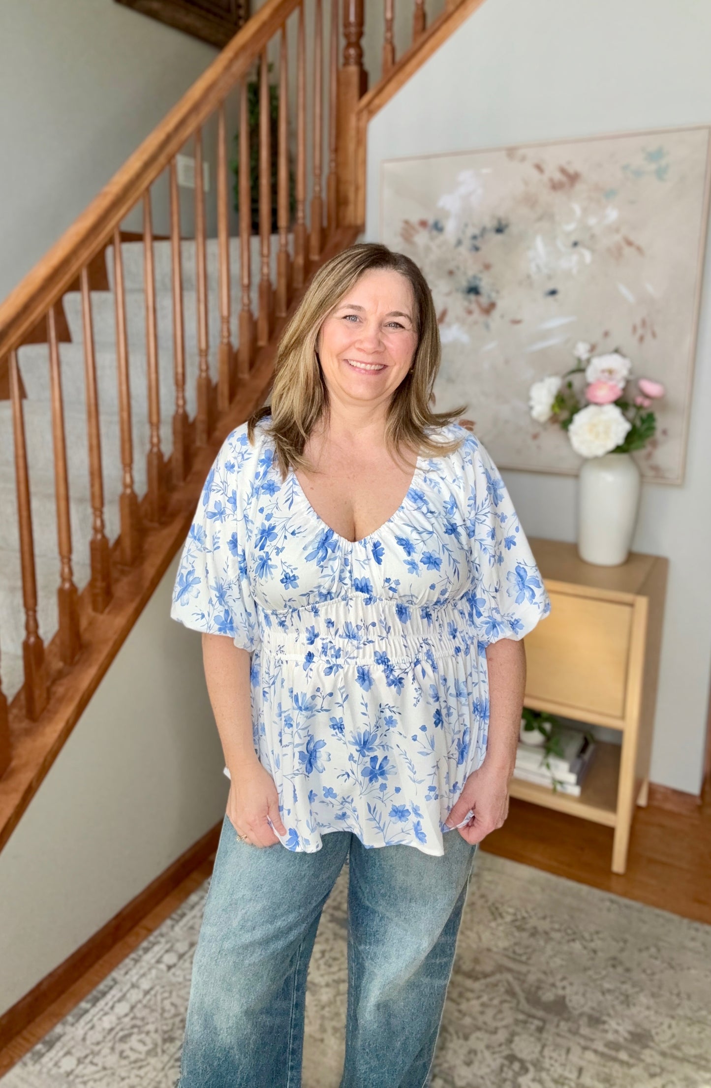 Woman wearing a blue floral blouse and jeans standing in a home setting with a staircase and decorative items.