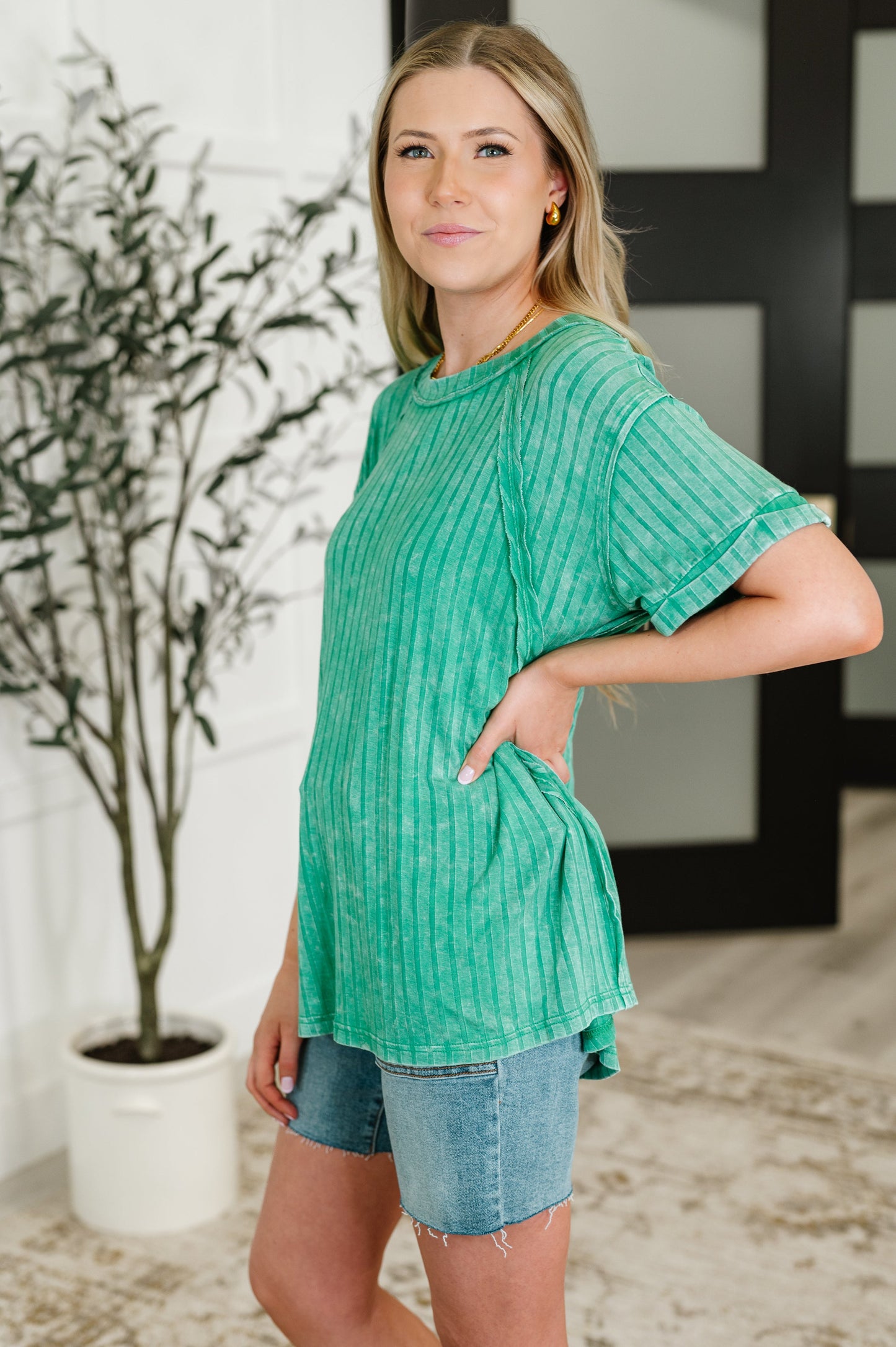 Woman wearing a green textured top and denim shorts standing indoors.