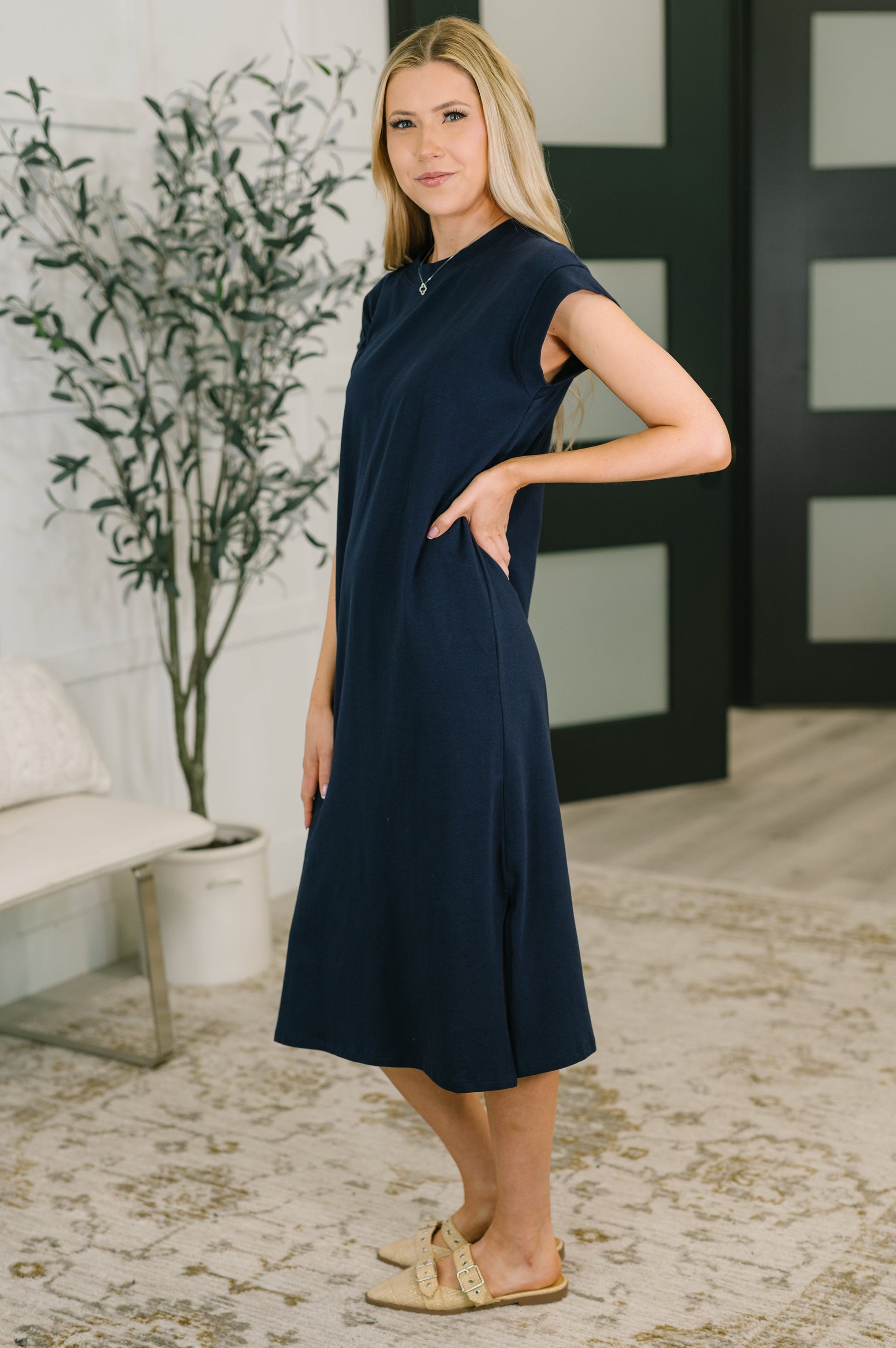 Woman wearing a navy dress standing in a room with a plant and furniture.