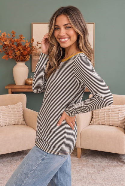 A woman modeling a long-sleeve striped top with a mustard yellow crew neck, paired with light blue jeans, in a cozy living room setting.
