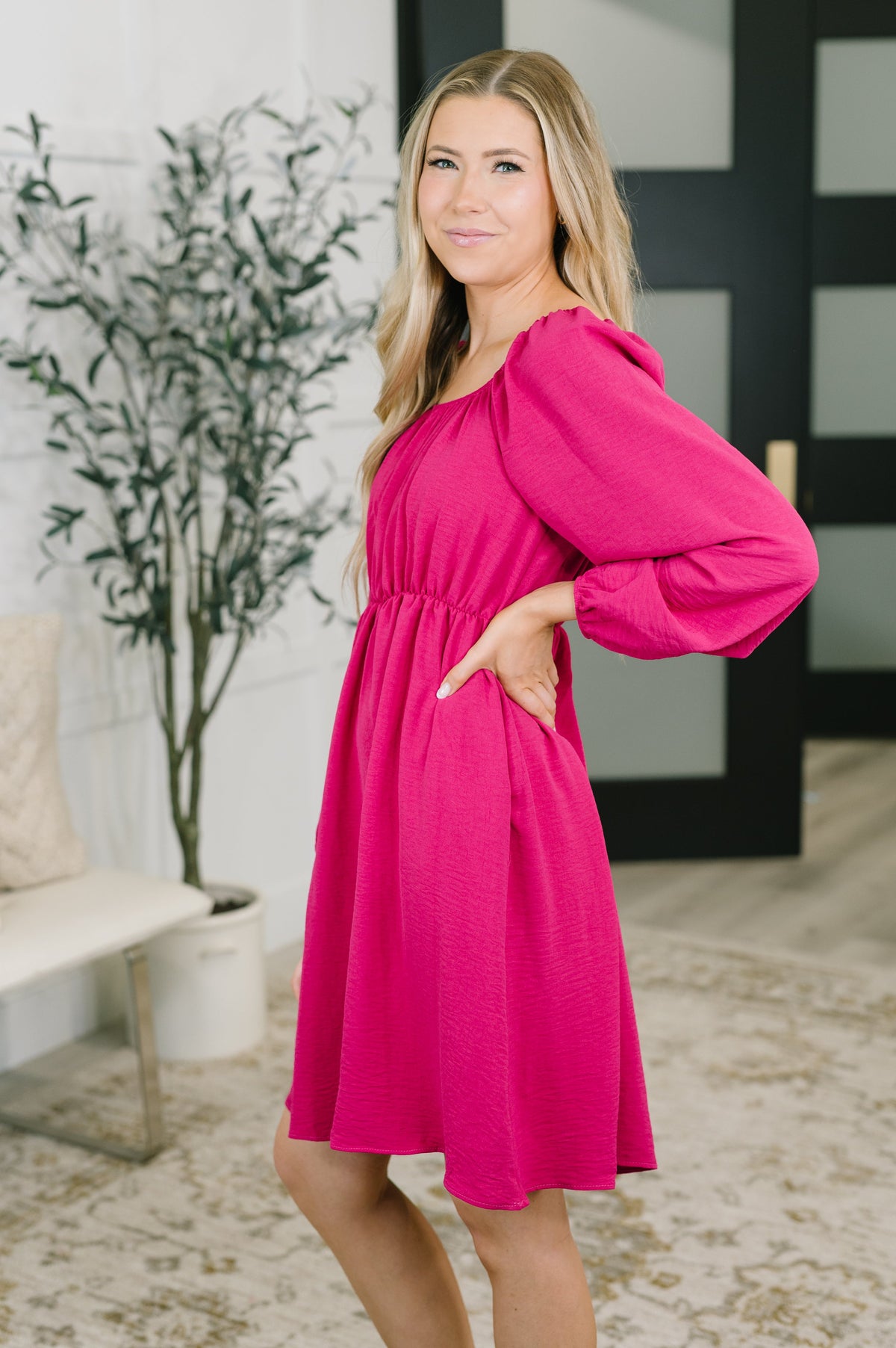 A woman modeling a bright pink puff-sleeve dress with a gathered waist and side pockets, standing in a modern indoor setting with a potted plant and light-colored floor.