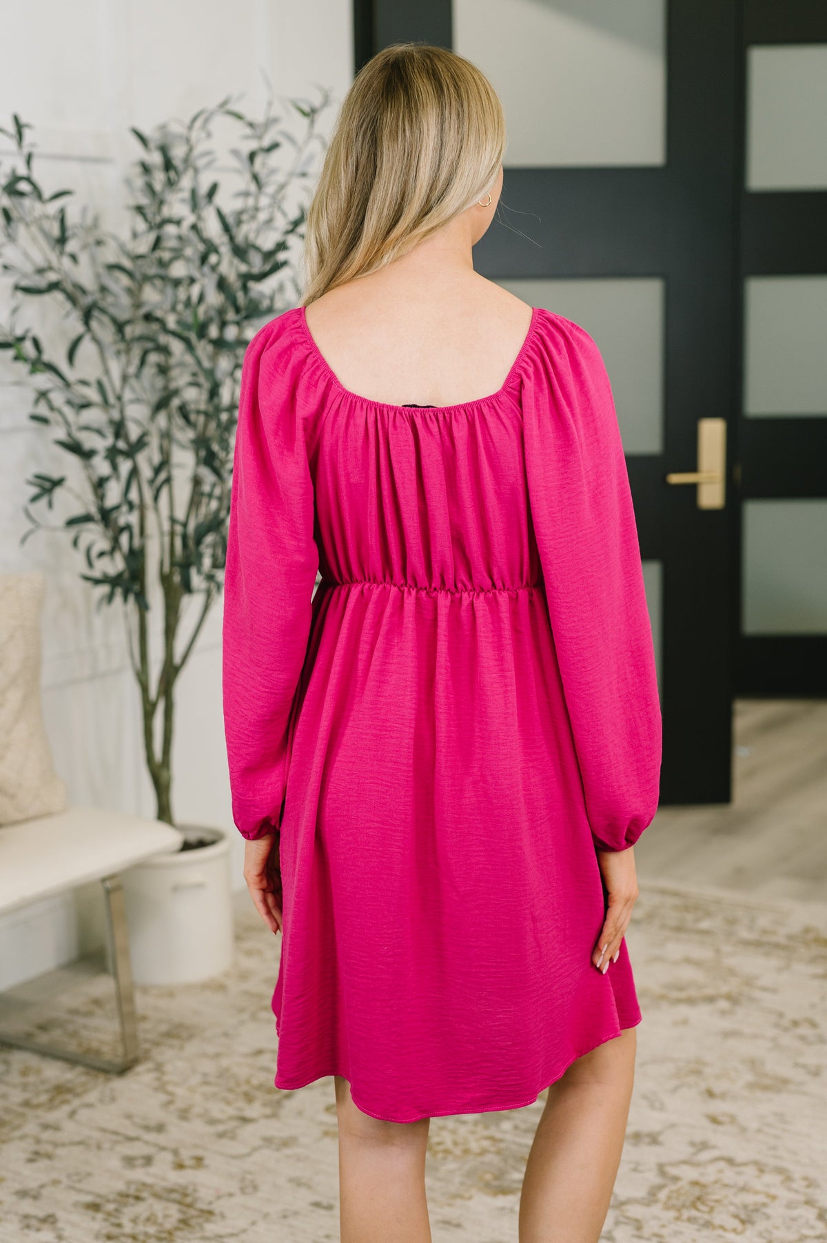 A woman viewed from behind wearing a bright fuchsia pink long-sleeve dress with a gathered empire waist and square neckline, standing in a modern indoor room with a plant and dark door in the background.