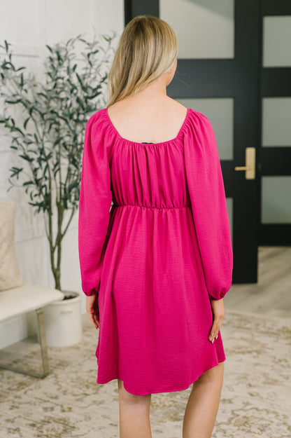 A woman viewed from behind wearing a bright fuchsia pink long-sleeve dress with a gathered empire waist and square neckline, standing in a modern indoor room with a plant and dark door in the background.