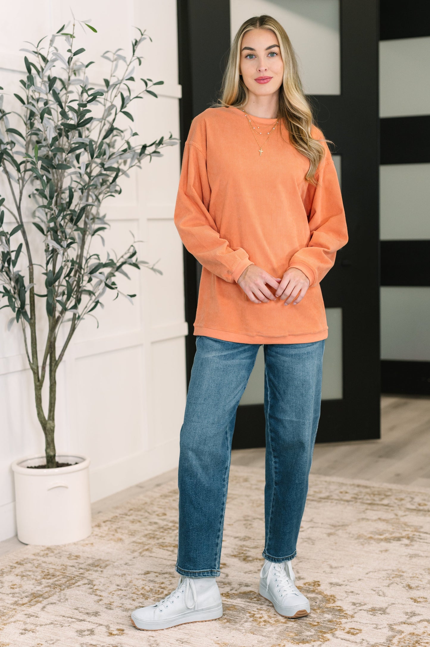 Woman wearing an orange sweater and blue jeans standing indoors with a plant in the background