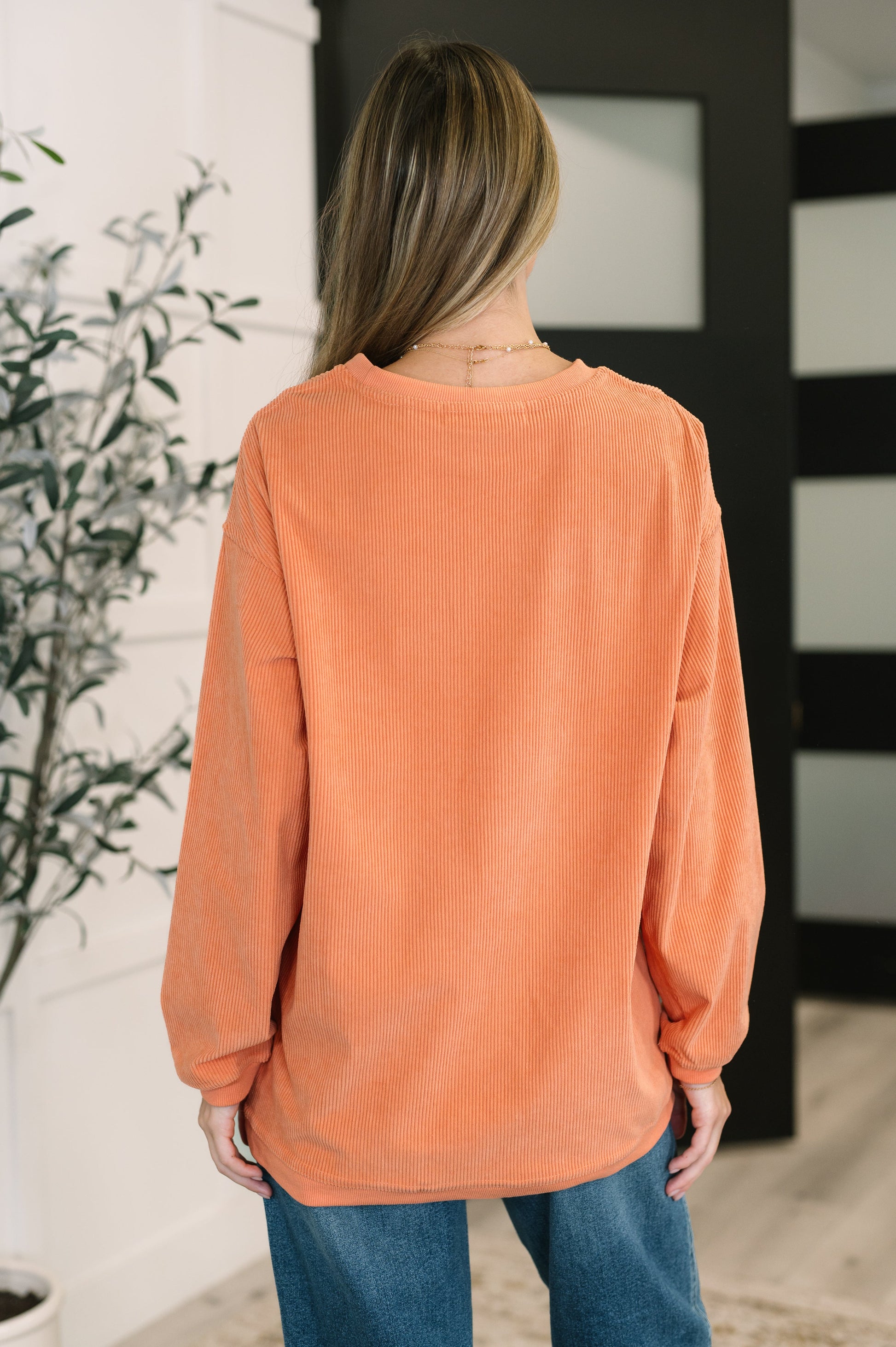 Person wearing an orange corded sweatshirt and blue jeans in a room with a plant and mirror.