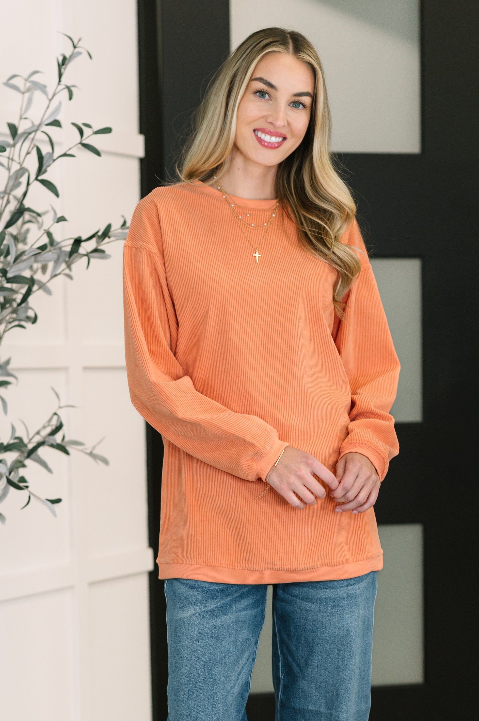 Woman wearing an orange corded long sleeve sweatshirt and blue jeans standing indoors.