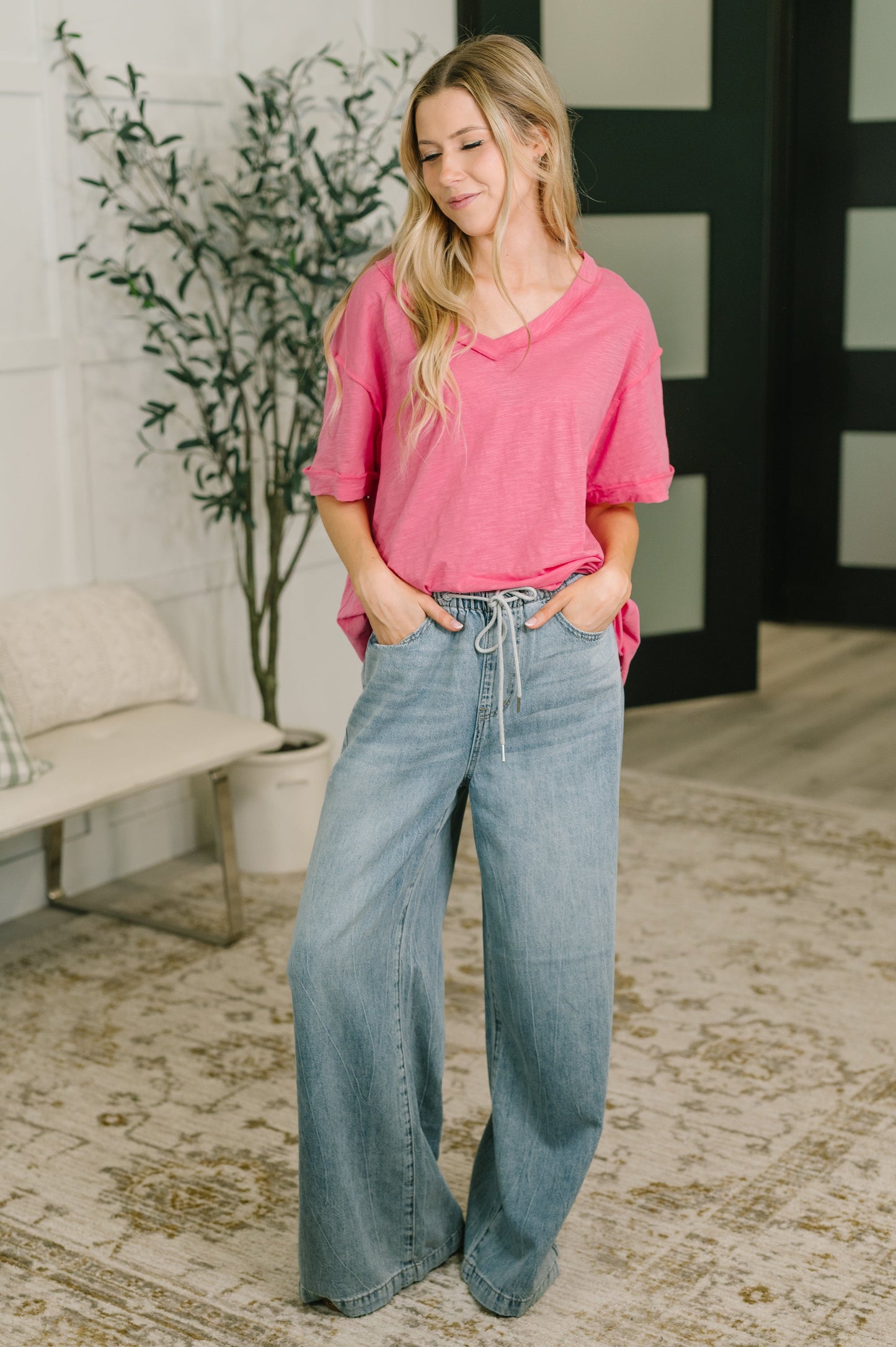 Woman wearing a pink top and blue jeans standing in a room with a plant and bench.