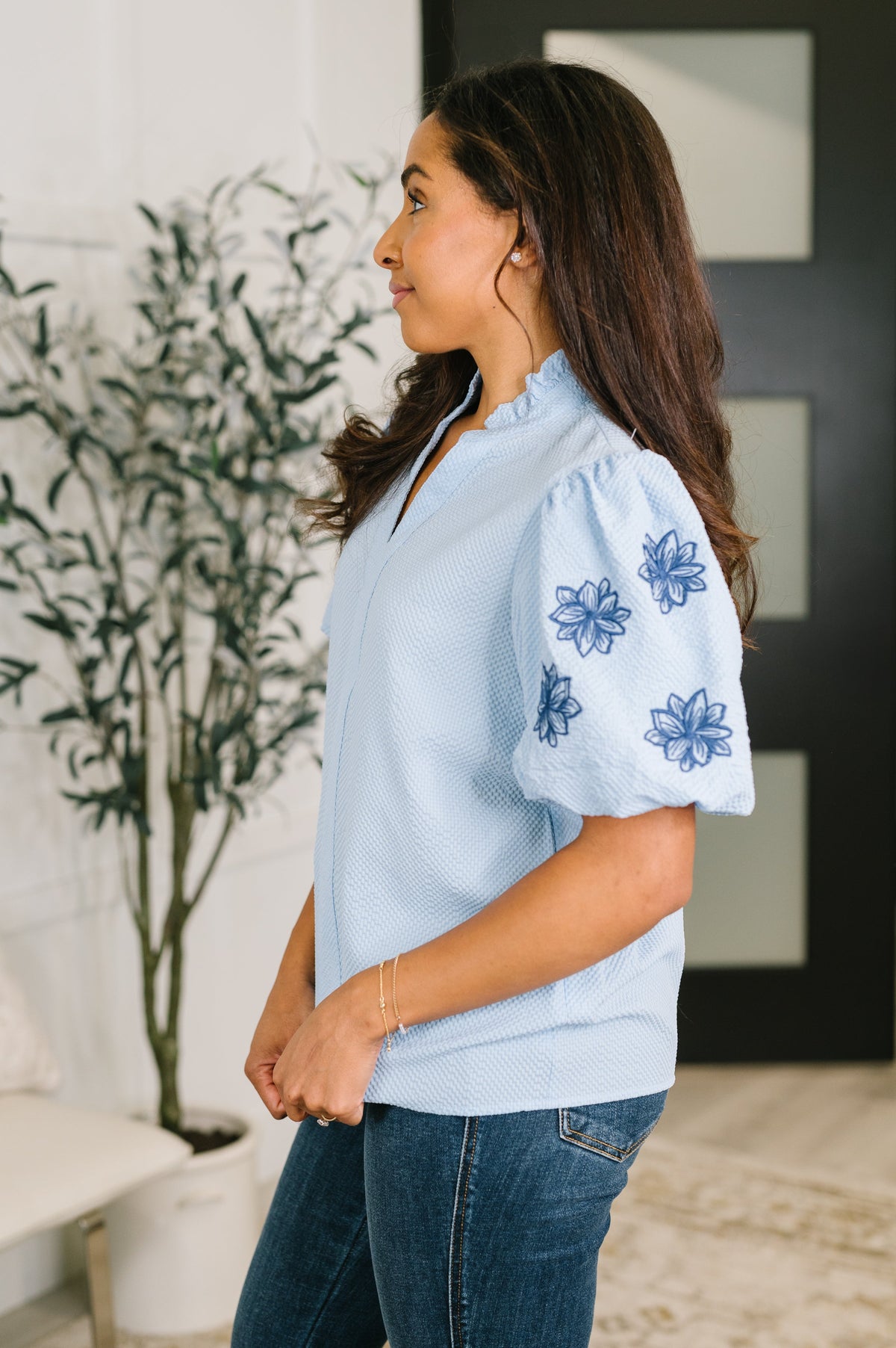 A woman wearing a light blue knit top with blue floral embroidery on the puff sleeves, paired with dark blue jeans, standing in a modern indoor setting.