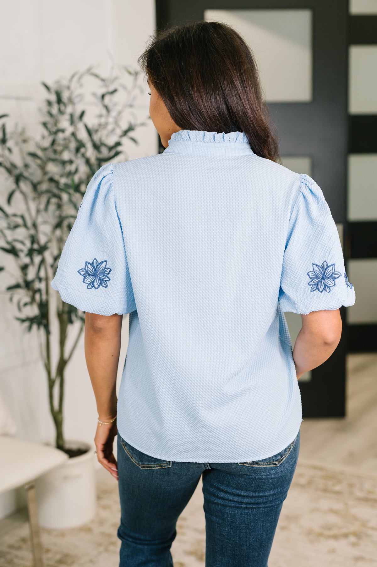 A woman viewed from behind wearing a light blue short-sleeve blouse with blue embroidered floral details on the sleeves, paired with dark blue jeans in an indoor setting.