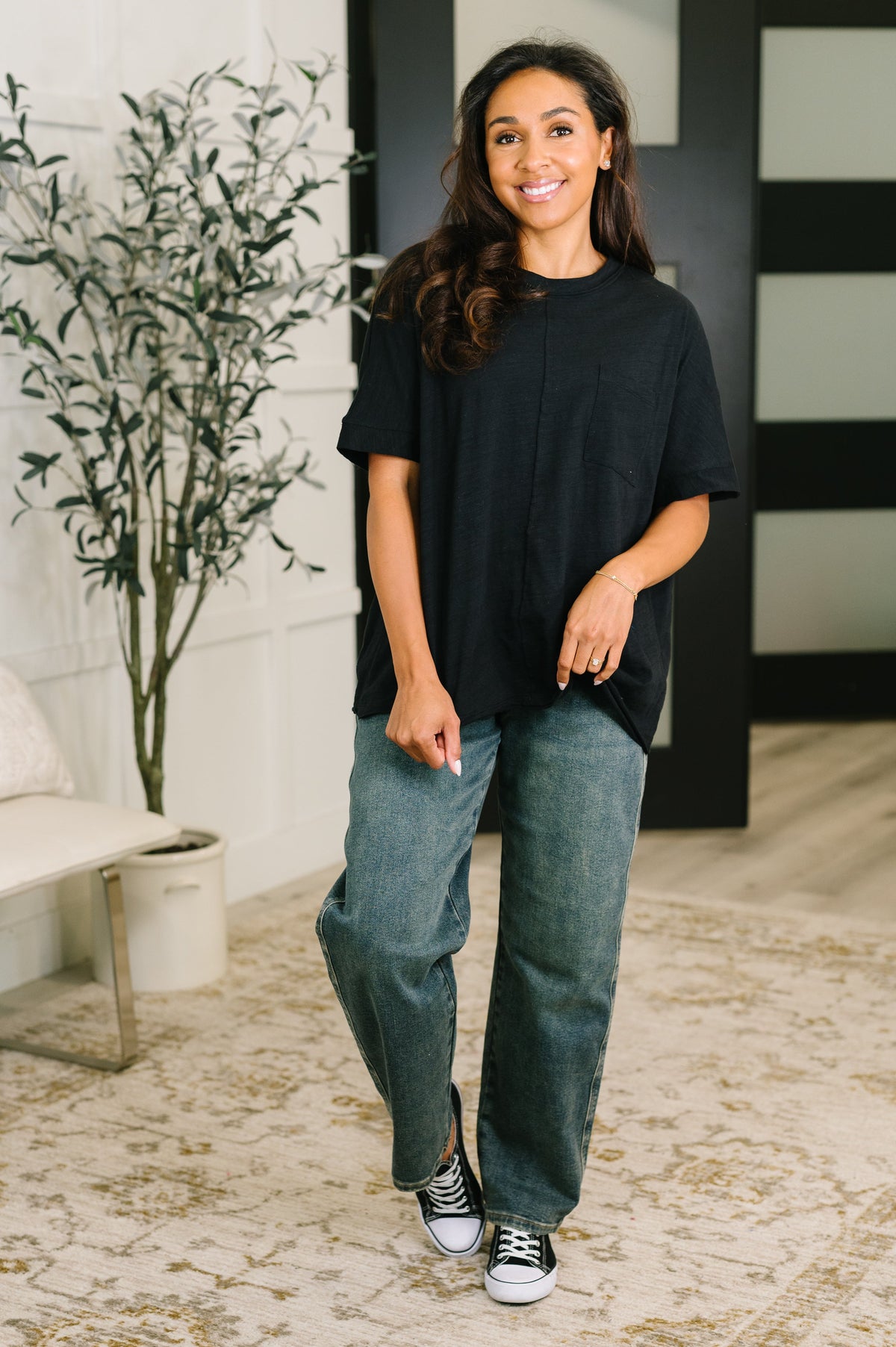 A woman modeling a black oversized short-sleeve t-shirt and wide-leg blue jeans, standing in a modern indoor room with a patterned rug, a potted plant, and a bench.