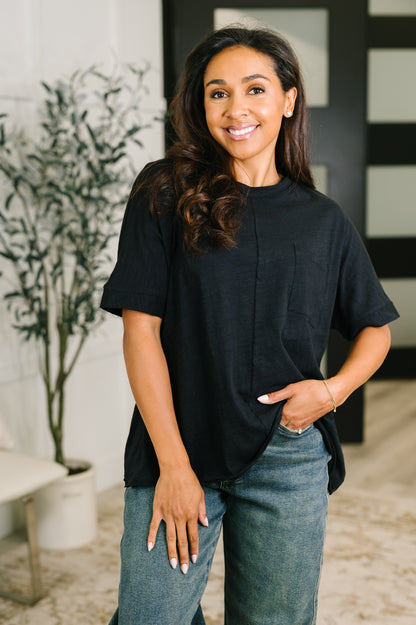 A woman wearing a black short-sleeve top and blue jeans, standing in a modern indoor setting with a potted plant and a black-and-white paneled wall in the background.