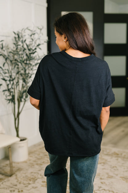 A woman viewed from behind wearing a loose-fitting black short-sleeve t-shirt and blue jeans in an indoor setting with a potted plant and a modern black door in the background.