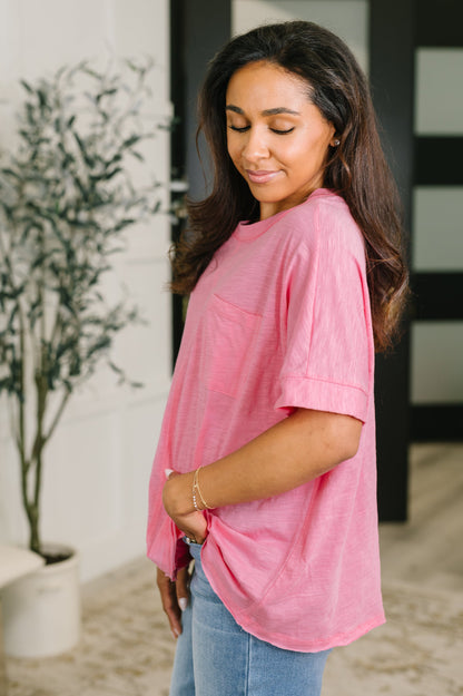 A woman wearing a pink short-sleeve crewneck top with a chest pocket, paired with light blue jeans, standing in an indoor setting.