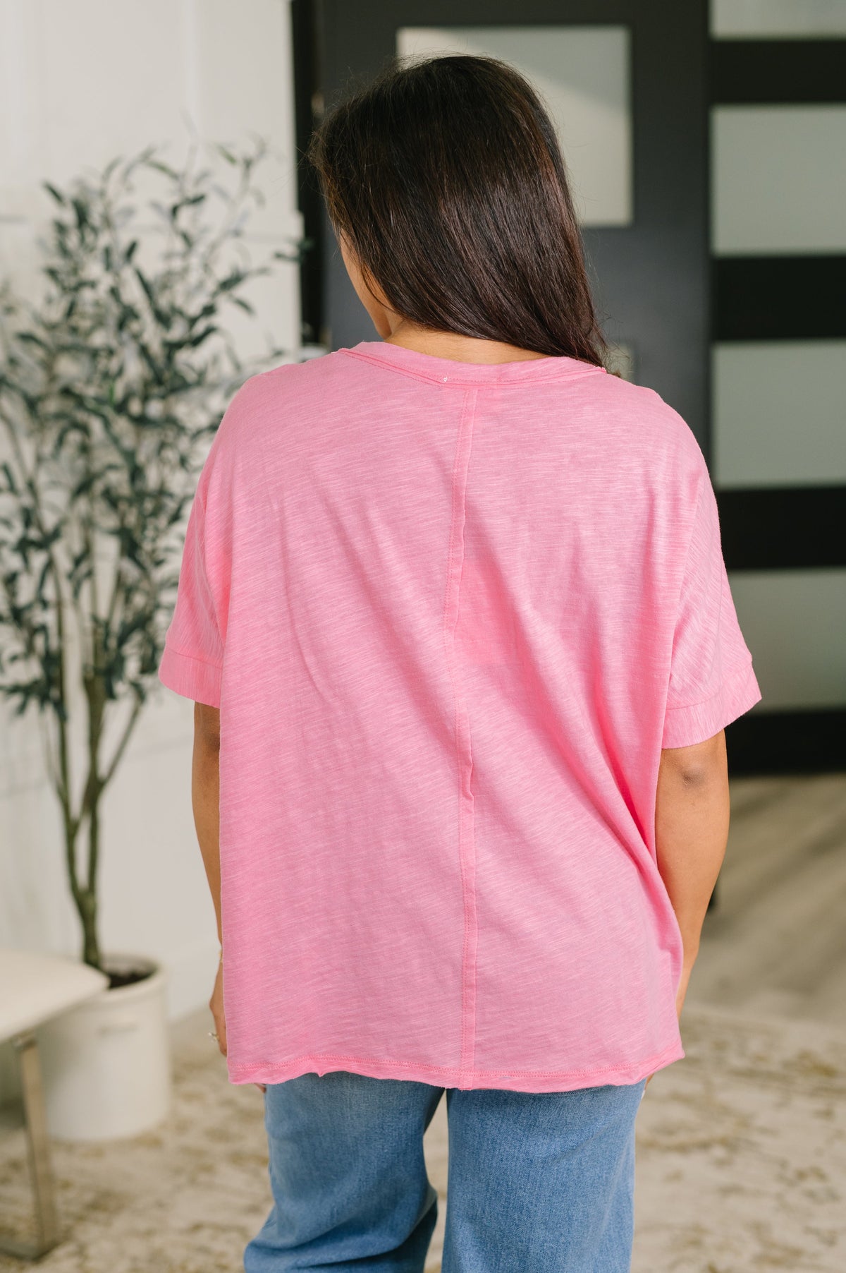 A woman viewed from behind wearing a loose-fitting pink short-sleeve t-shirt and blue jeans in an indoor setting.