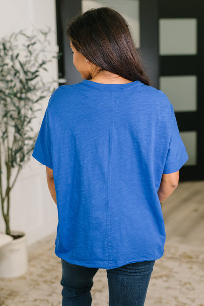 A woman viewed from behind wearing a bright blue short-sleeve t-shirt and dark blue jeans in an indoor setting.