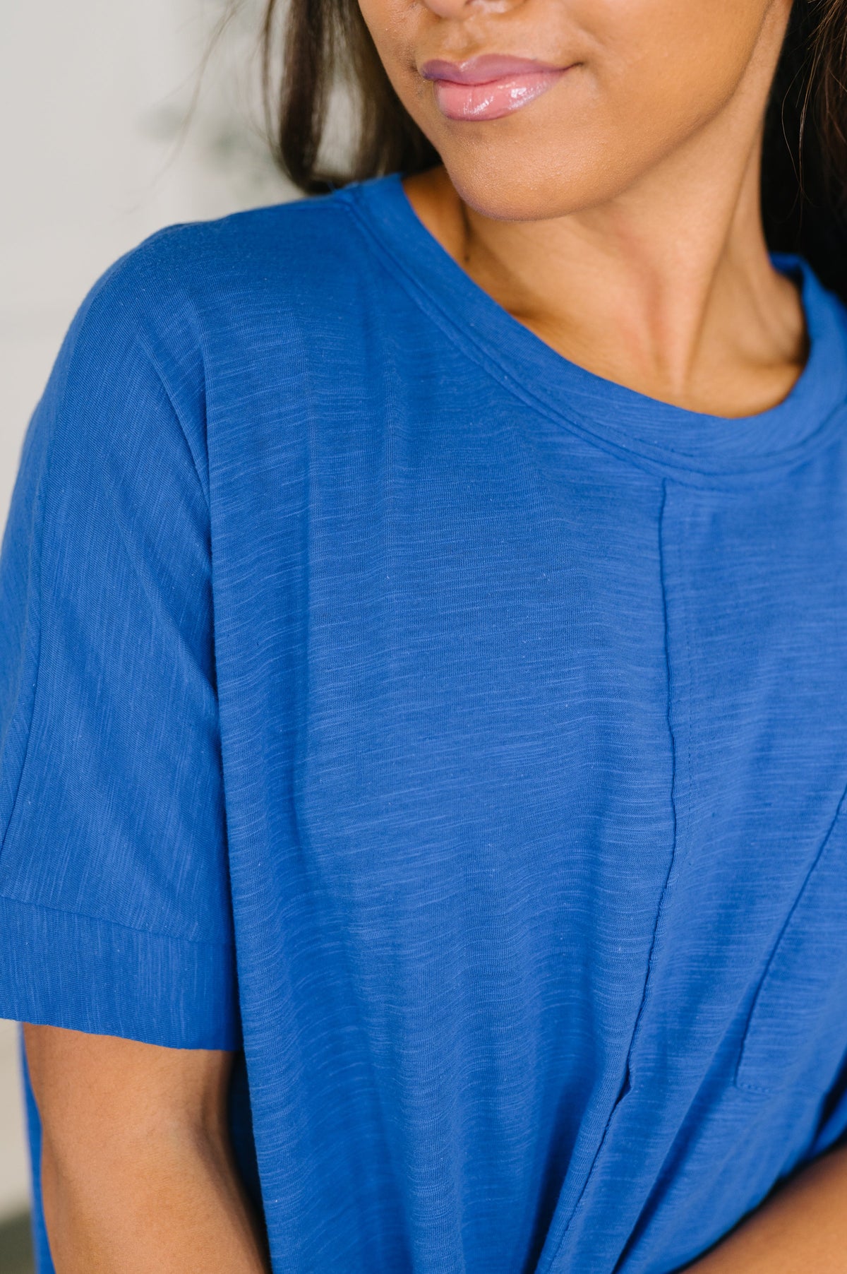A close-up of a person wearing a short-sleeve crew-neck top in a medium blue color, showing the fabric texture and a vertical seam detail on the front.