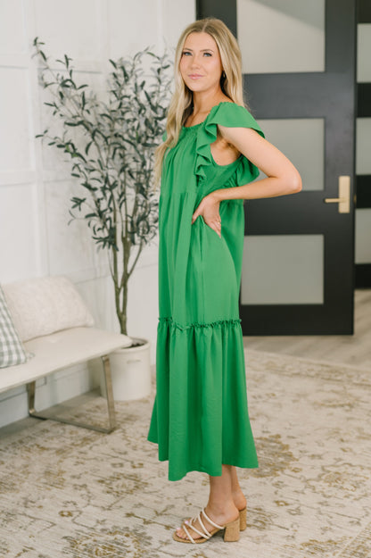 Woman wearing a green dress standing in a room with a plant and door in the background.