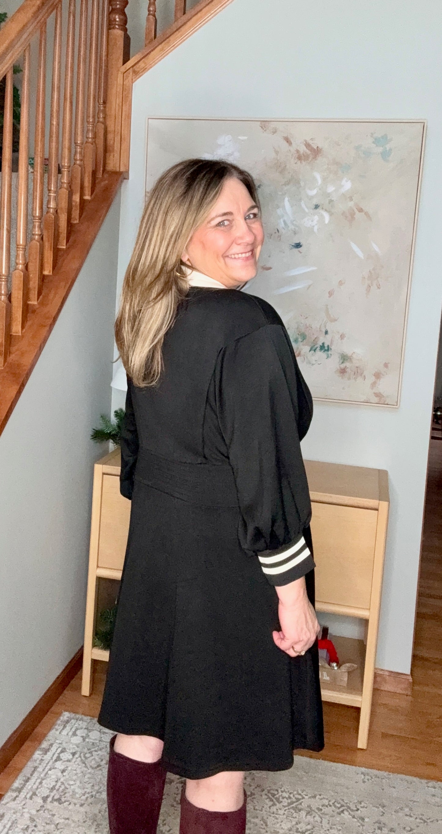 Woman wearing a black dress with white stripes on the sleeves, standing in a room with wooden stairs and a painting on the wall.