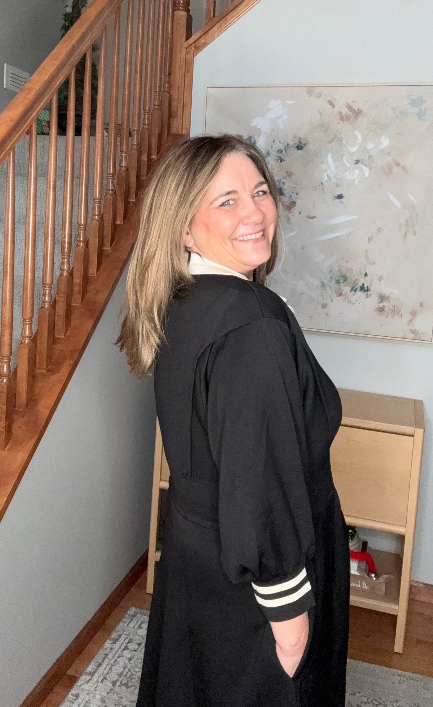 Woman in a black outfit with white stripes standing in a home setting with a staircase and artwork on the wall.