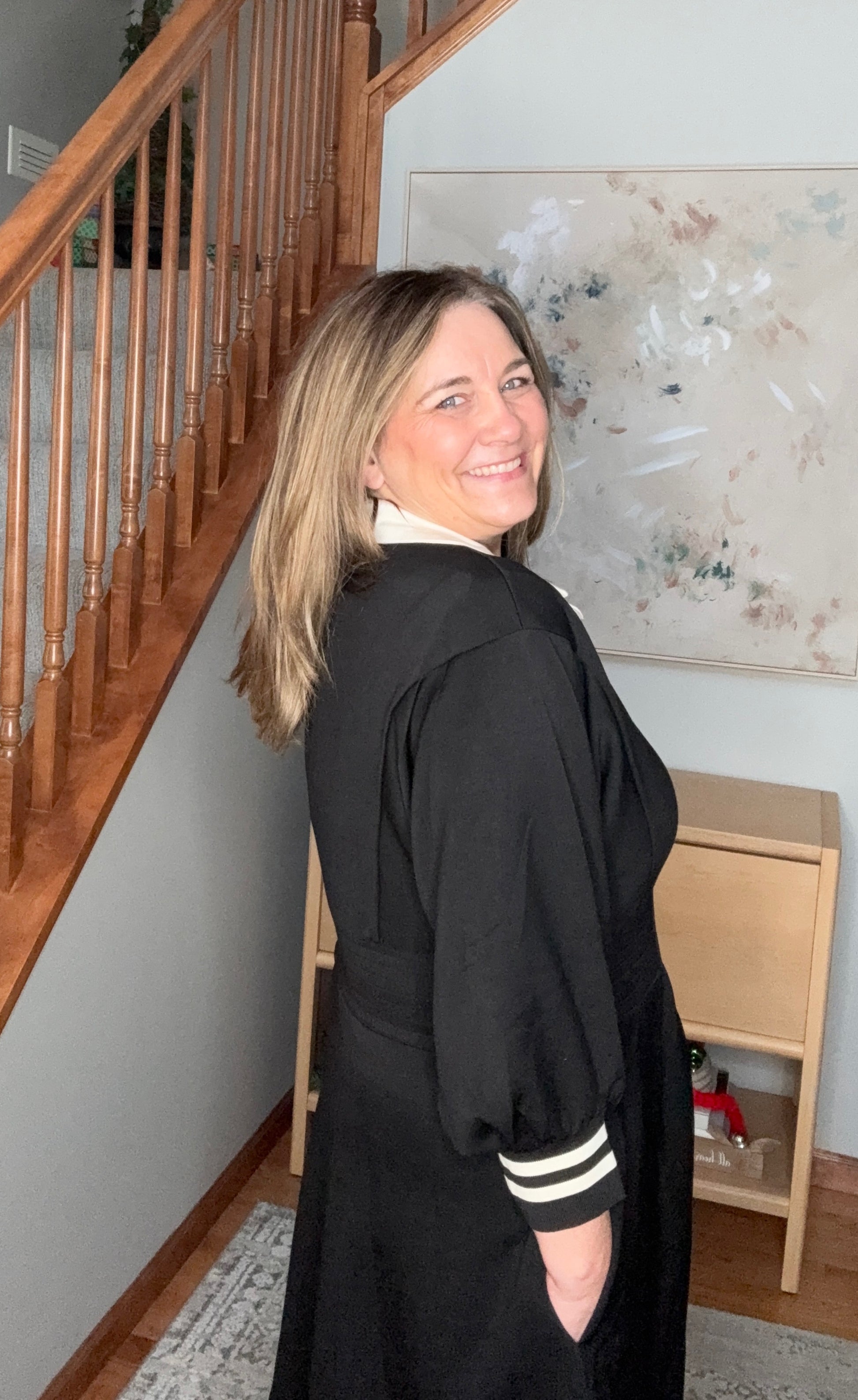 Woman in a black outfit with white stripes standing in a home setting with a staircase and artwork on the wall.