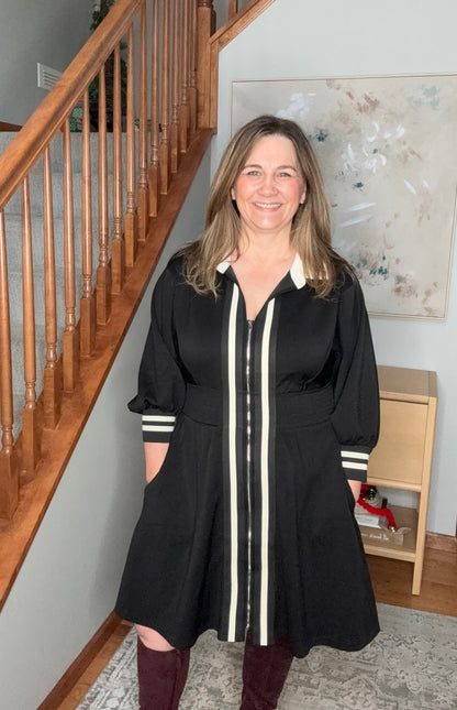Woman in a black dress with white stripes standing in a home setting with wooden staircase and decor.