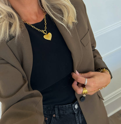 Person wearing a gold heart-shaped necklace, black top, and brown blazer with a neutral background.