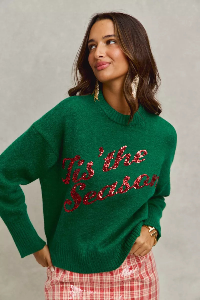 Woman wearing a green sweater with red sequins saying tis the season, standing against a plain background