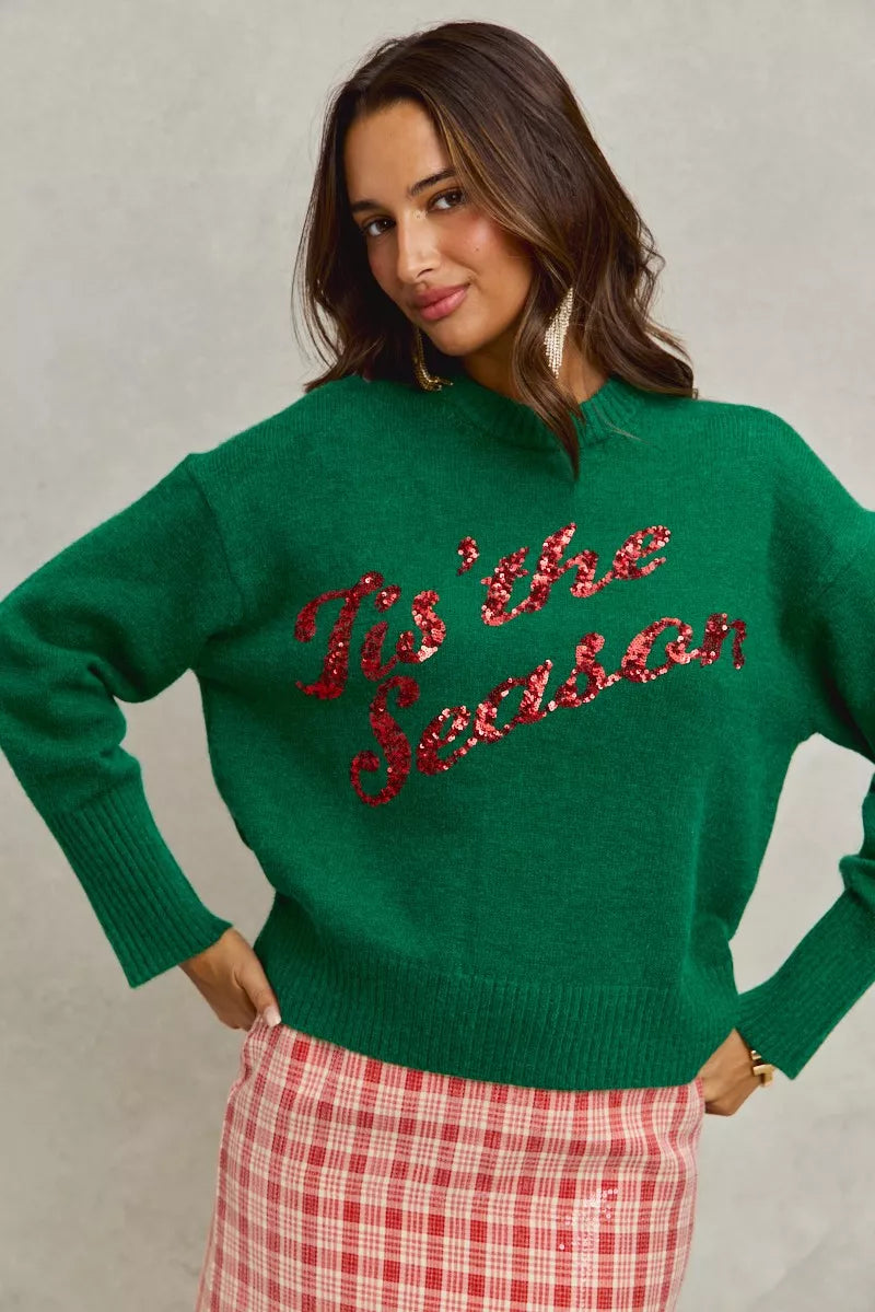 Woman wearing a green sweater with 'Tis the Season' text, standing against a plain background
